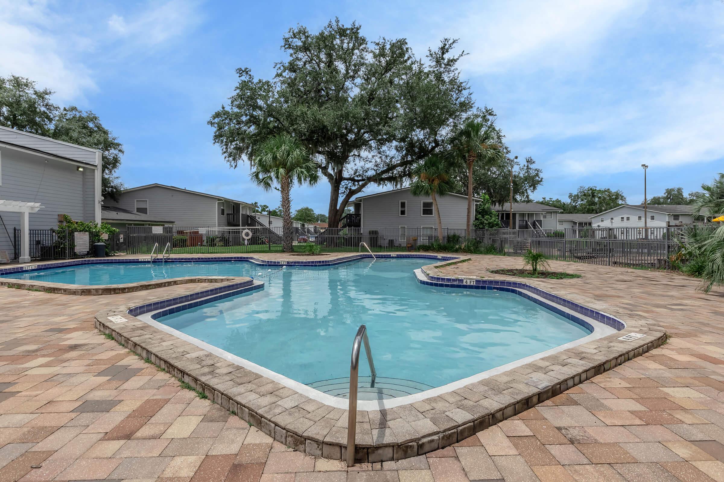 A clean, inviting swimming pool surrounded by a spacious patio made of stone pavers. Lush greenery and residential buildings are in the background, providing a tranquil atmosphere. The sky is clear with a few clouds, suggesting a pleasant day for swimming or relaxation.