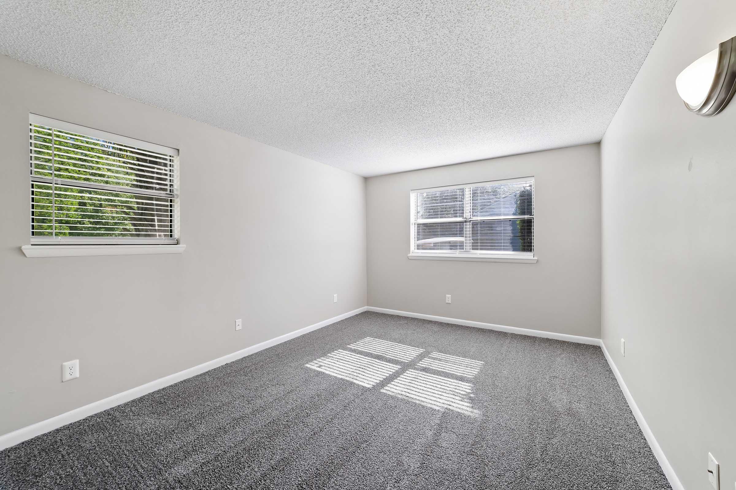Empty room with light gray walls and a textured ceiling. Two windows with closed blinds allow natural light to filter in, casting shadows on the light gray carpeted floor. The space is uncluttered, offering a clean and spacious feel, ideal for various interior design options.