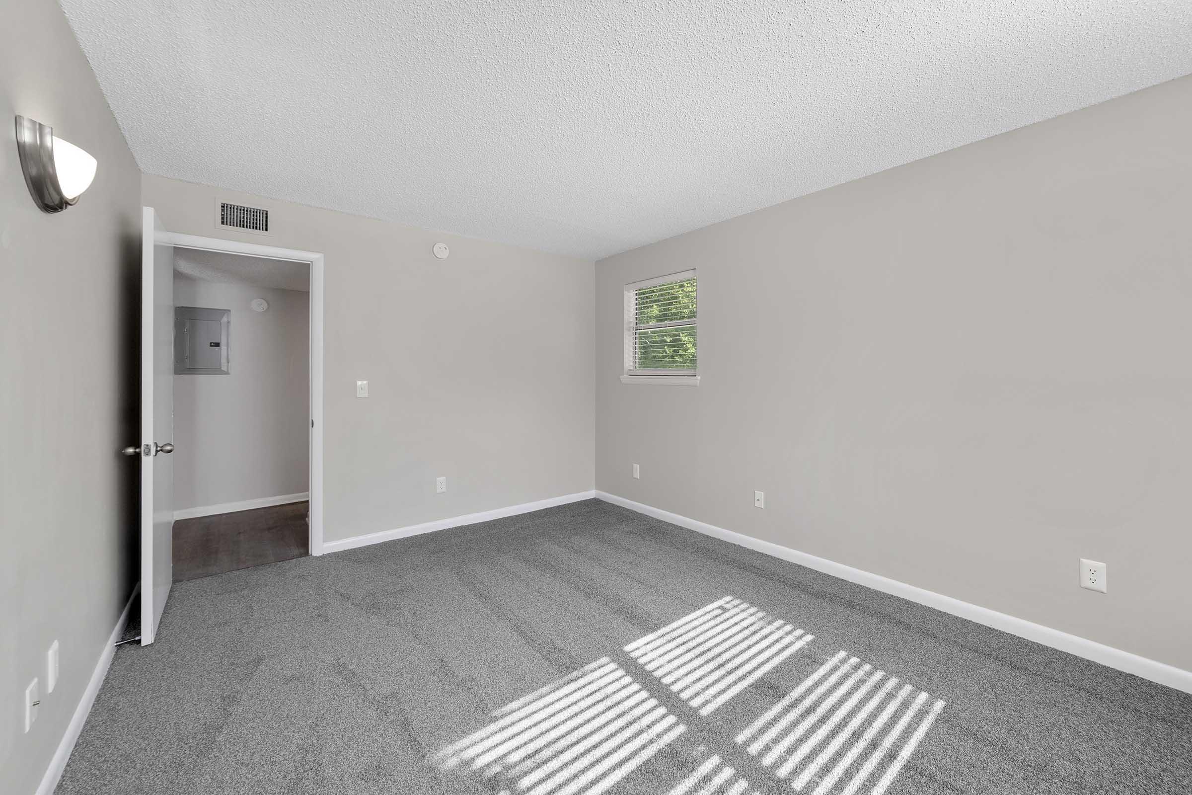 A light and airy room featuring gray carpeting, cream-colored walls, and a single window allowing natural light. The room has a simple design with a door leading to another space and a light fixture on the wall, giving it a clean and fresh appearance.