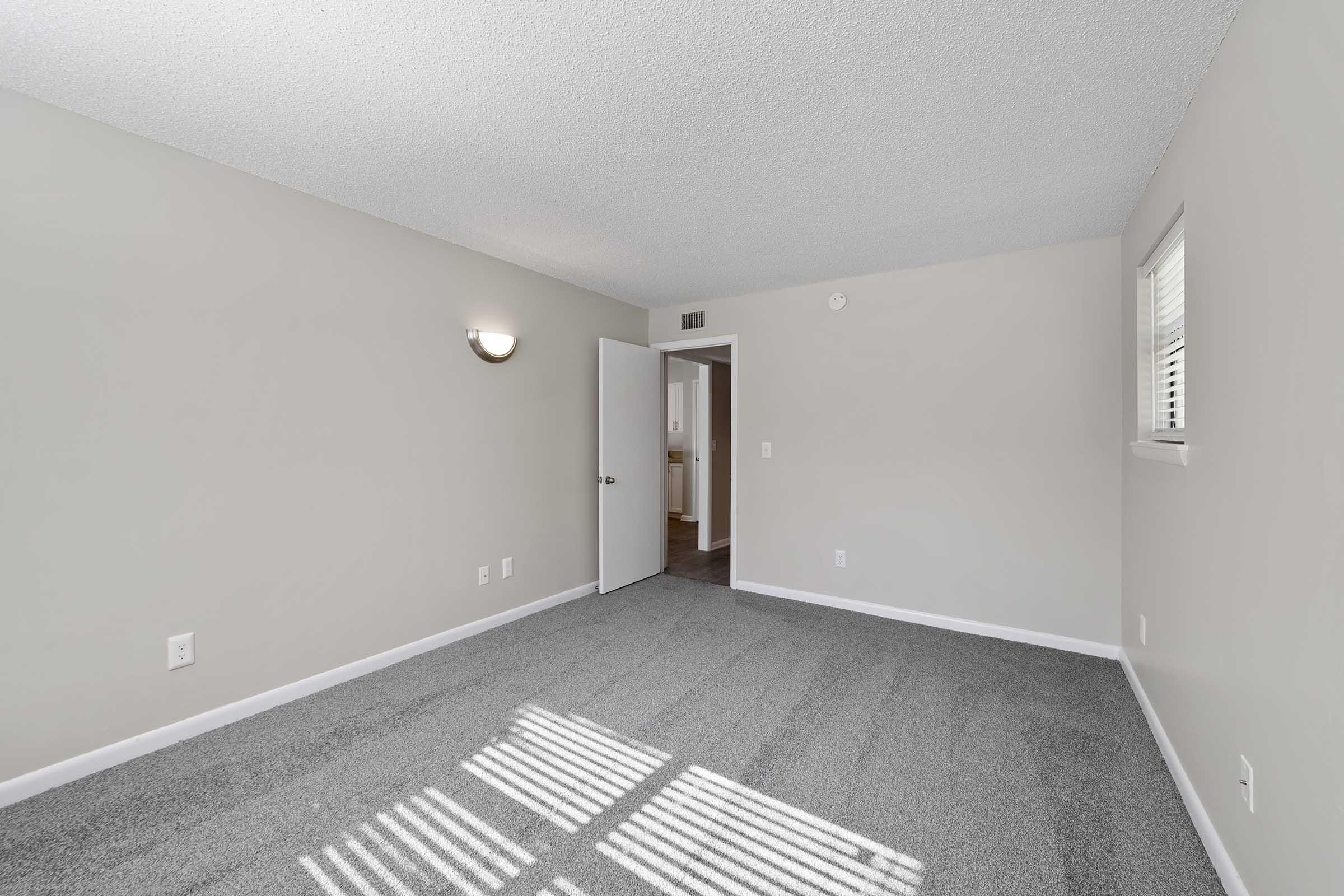 A well-lit, empty room featuring light gray carpet and neutral-colored walls. The room includes a wall-mounted light fixture and a white door leading to another space. Sunlight casts striped shadows on the floor from the window, creating a bright, open atmosphere.