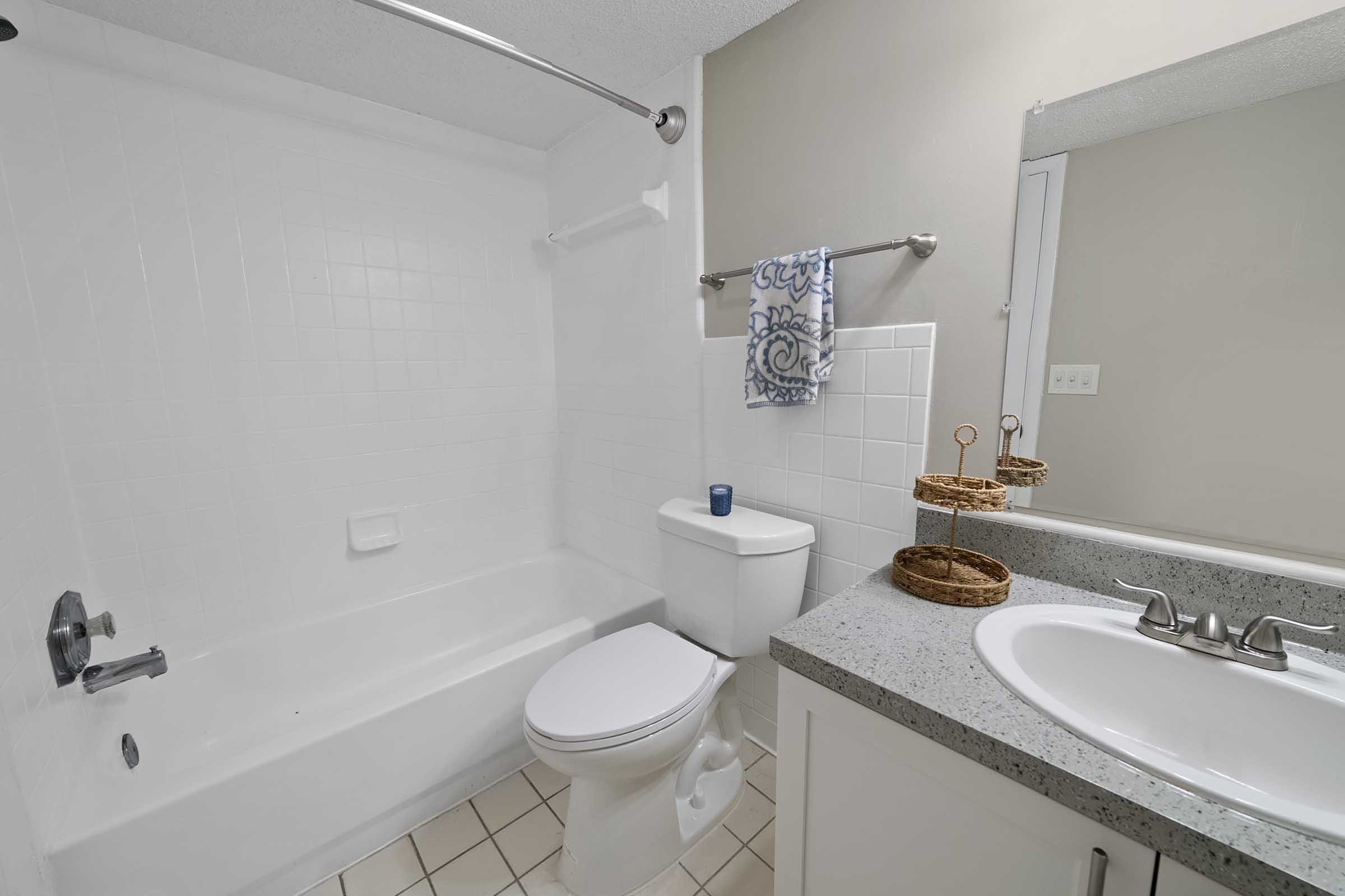 A clean and well-lit bathroom featuring a bathtub with a shower, a modern toilet, and a sink with a countertop. The walls are painted in a soft color, and there are decorative elements like a towel and woven baskets on the countertop. The overall design is simple and functional.