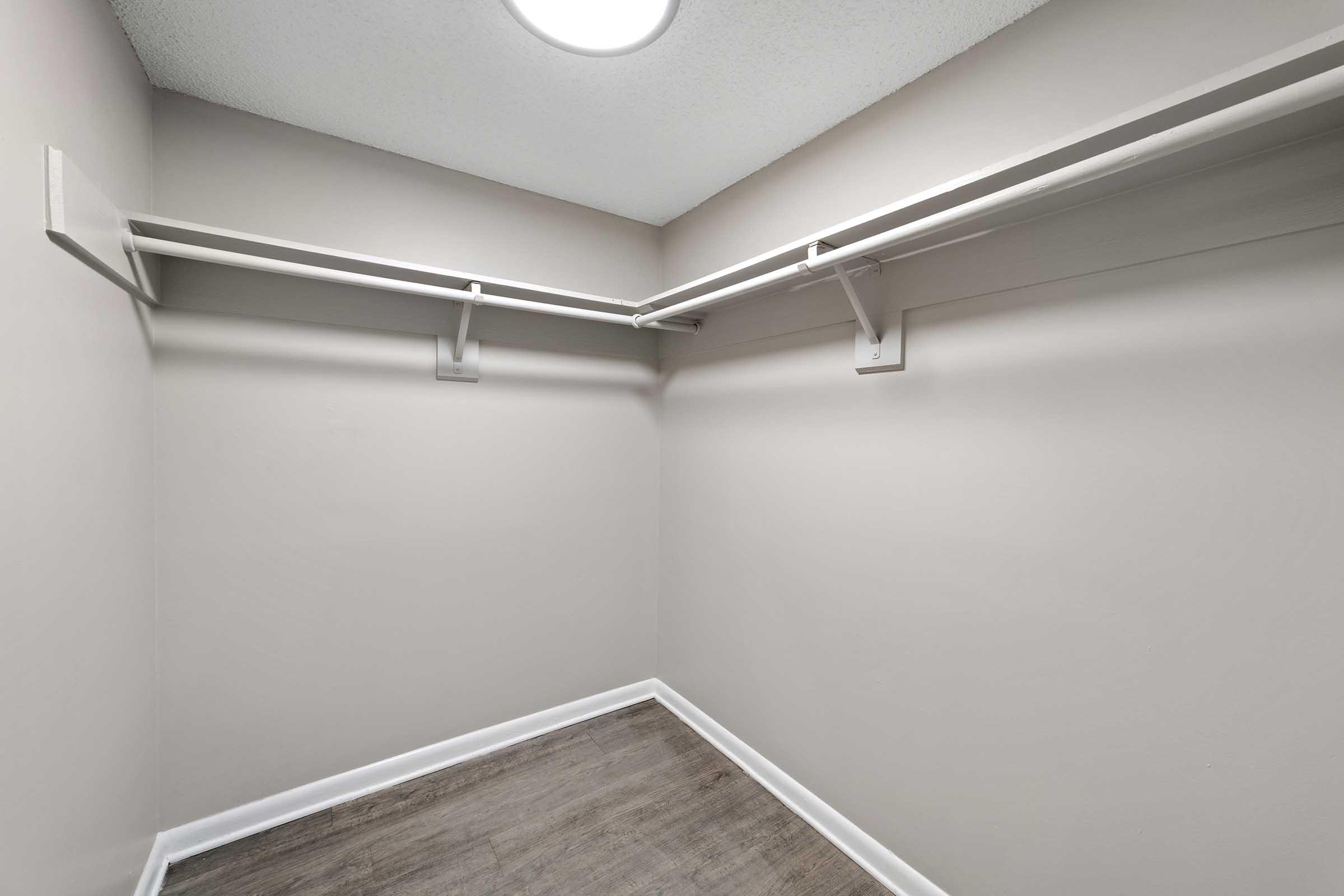 Empty closet with light gray walls and a laminate floor. It features two hanging rods for clothes, mounted on the wall. The ceiling has a circular light fixture. The overall space appears clean and organized, suitable for storage or as a dressing area.