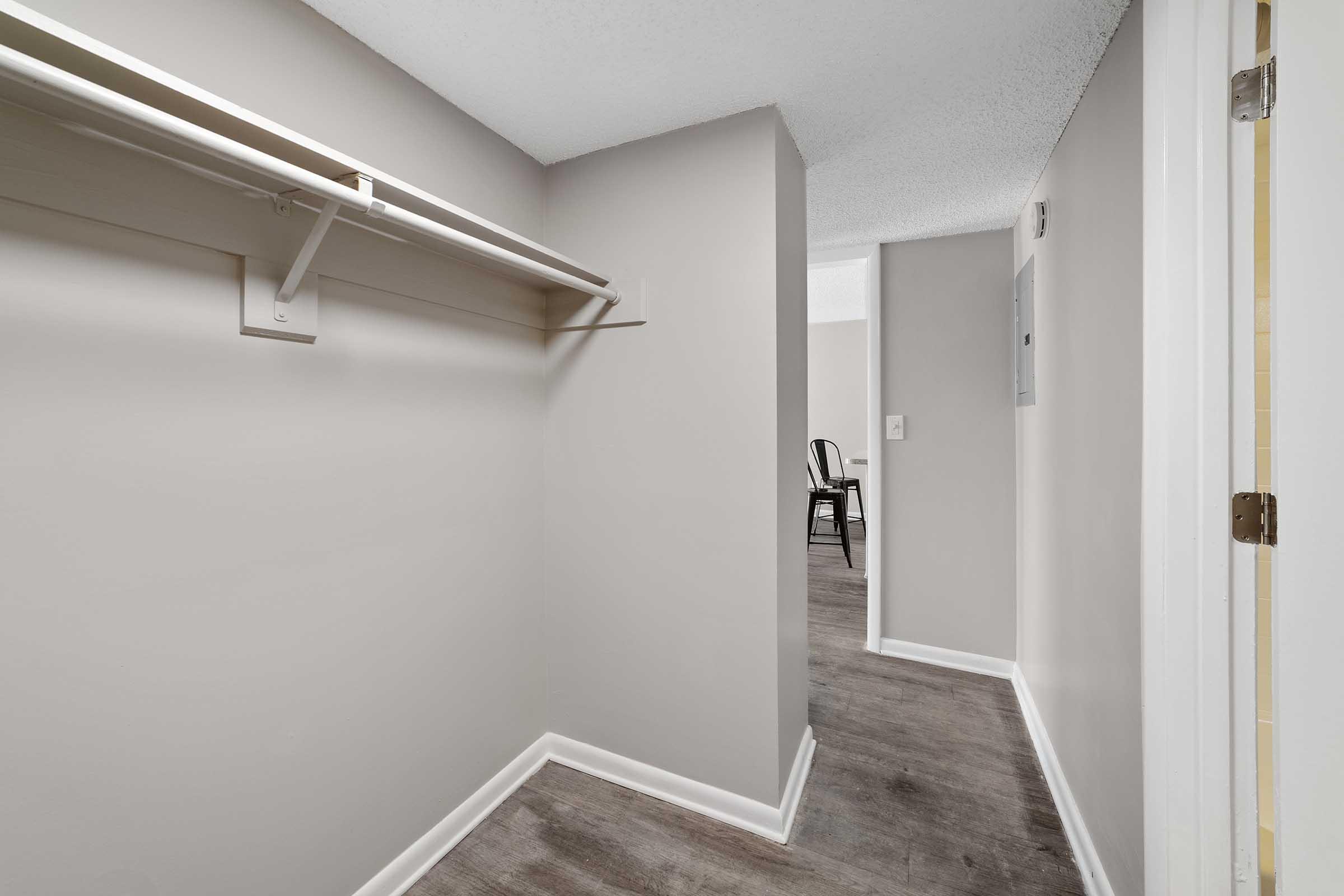 A narrow hallway in a lightly painted room, featuring a white shelving unit on the left. The floor is a wood-like material, and in the background, a glimpse of a chair and another room is visible, suggesting an open layout. The space is well-lit and appears tidy.