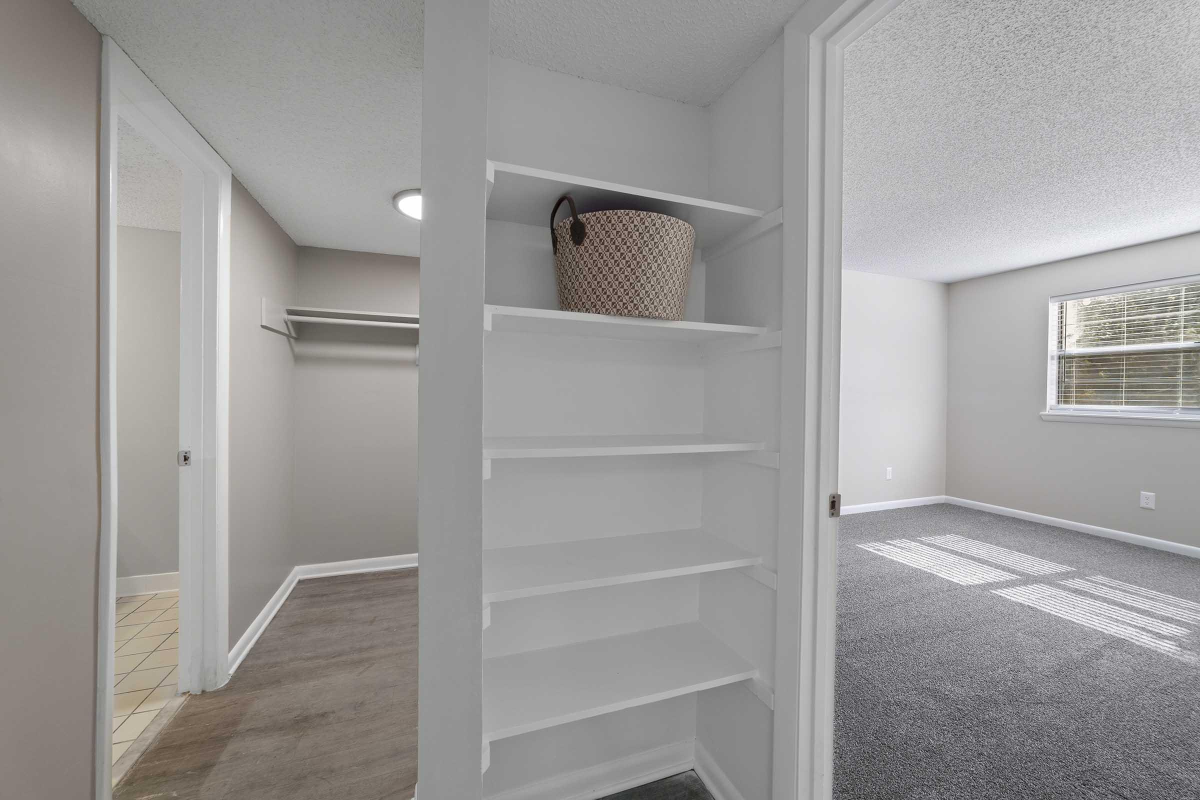A view of a light-colored hallway with a white shelving unit on the left. A basket sits on one of the shelves. To the right, there is a carpeted room with a window allowing natural light, and a closet space visible. The walls are painted in neutral tones, creating a spacious feel.