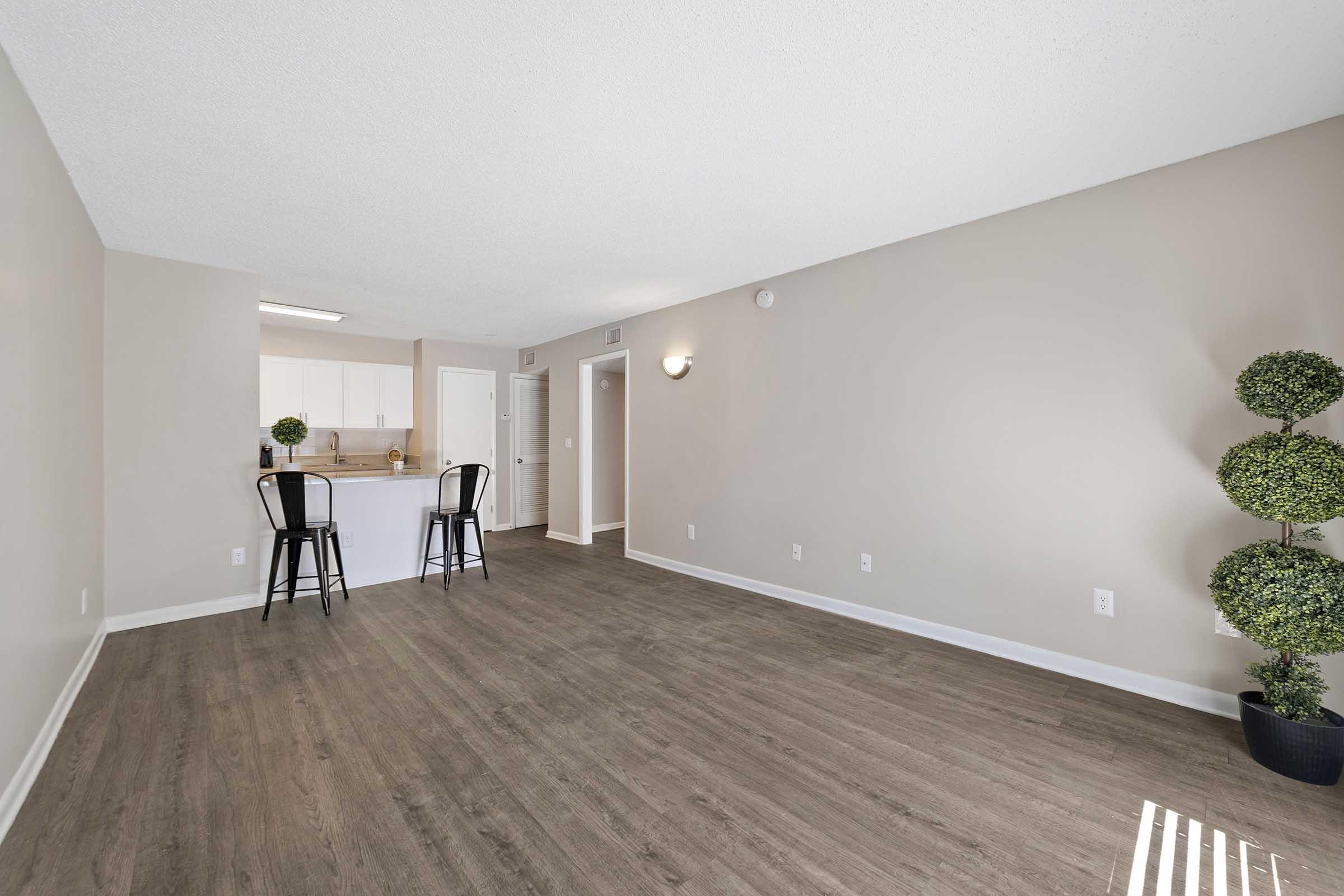 Spacious, empty living area with hardwood-style flooring, light beige walls, and subtle lighting. In the background, a kitchen area with bar stools is visible, along with a doorway. Two decorative topiary plants are placed in the corner, adding a touch of greenery to the modern space.