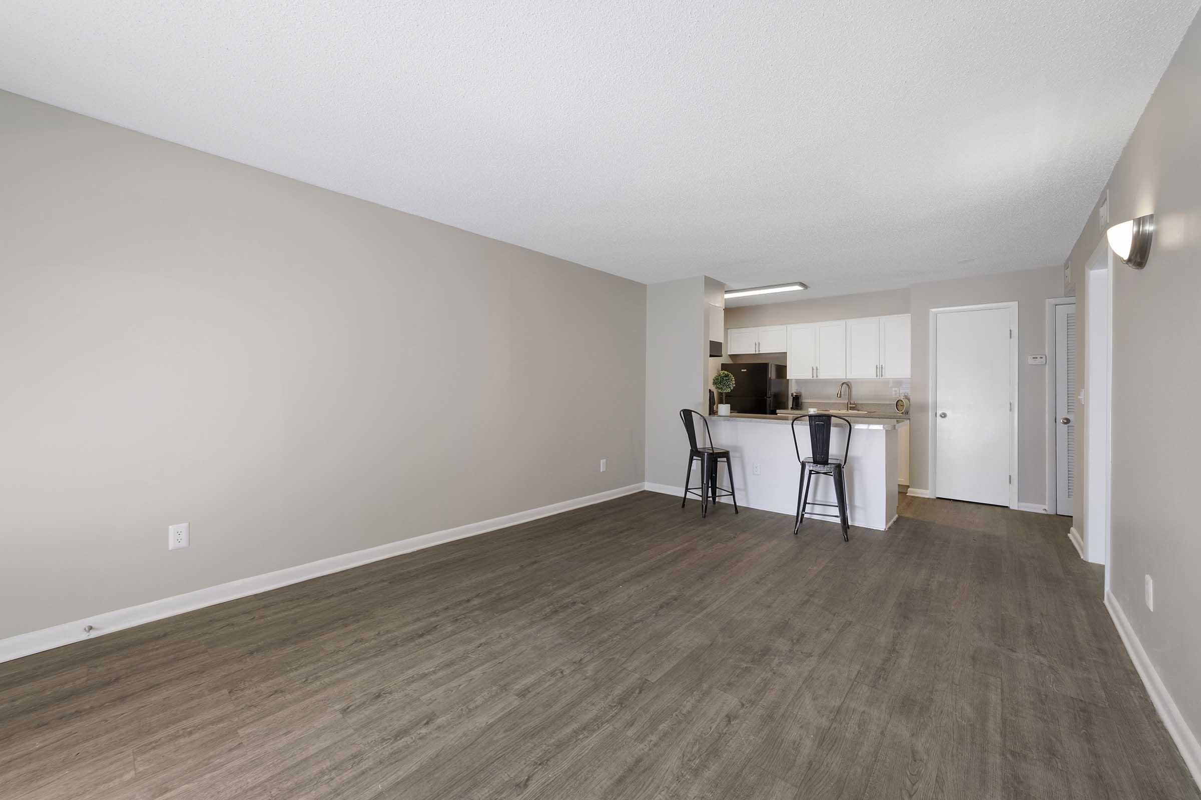 Spacious interior of a modern apartment featuring a neutral color palette, hardwood-style flooring, and a kitchen with a bar counter. Two black stools are positioned at the counter, creating a cozy dining space. The layout is open, with natural light enhancing the atmosphere.