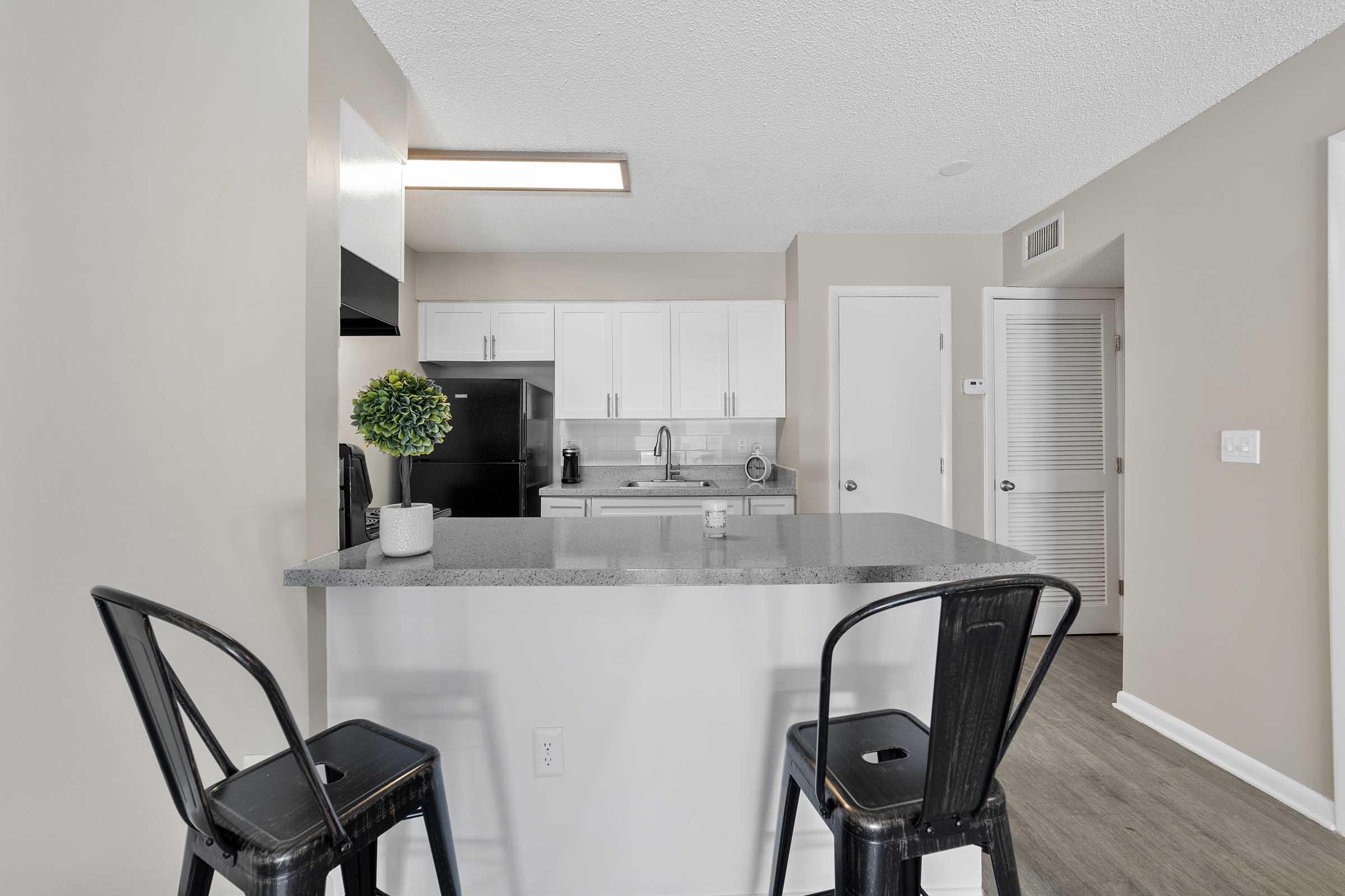 A modern kitchen with a gray countertop and white cabinetry. Two black bar stools are positioned at the countertop. The kitchen features a black refrigerator, and there's a small plant on the countertop. The room has light-colored walls and a door leading to another area.