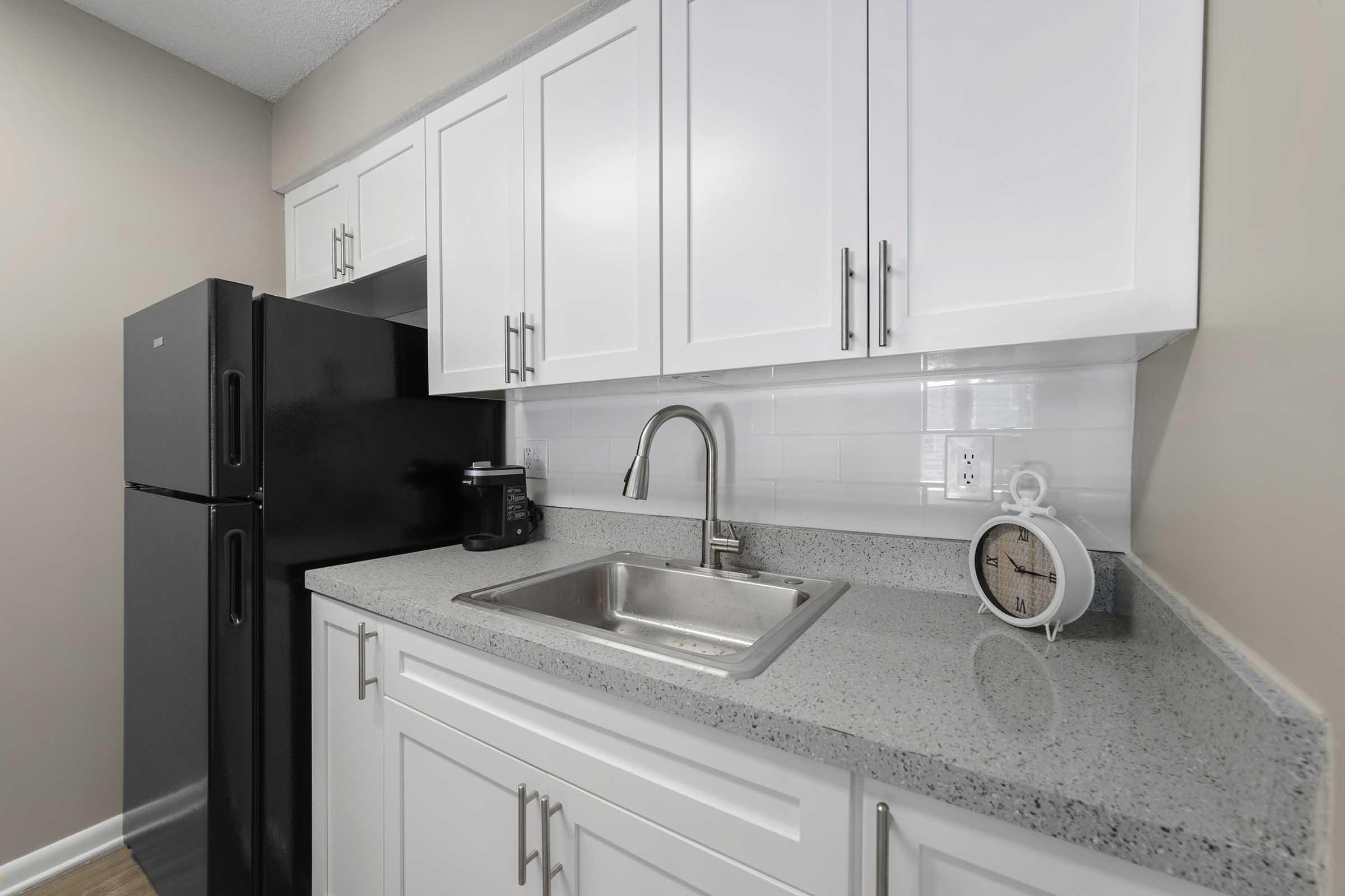 Modern kitchen with white cabinets, a stainless steel sink, and a black refrigerator. There is a small clock on the countertop, and the backsplash features white tiles. The overall design is sleek and contemporary, with soft lighting.
