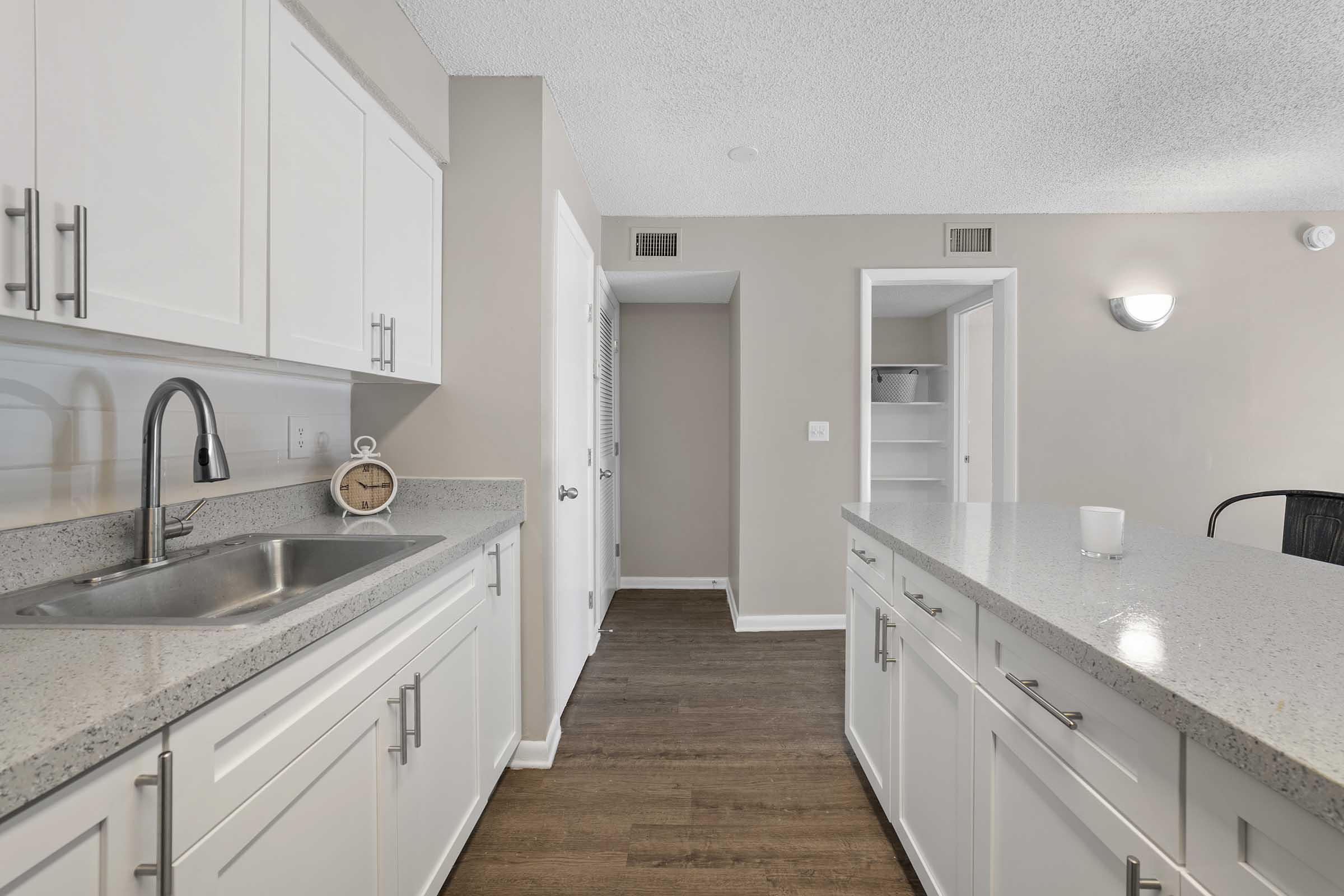Modern kitchen interior featuring white cabinetry, a granite countertop with a sink, and a small decorative clock. The space has a neutral wall color and a door leading to another room, creating an open and inviting atmosphere. A chair is visible in the background, adding a homey touch.