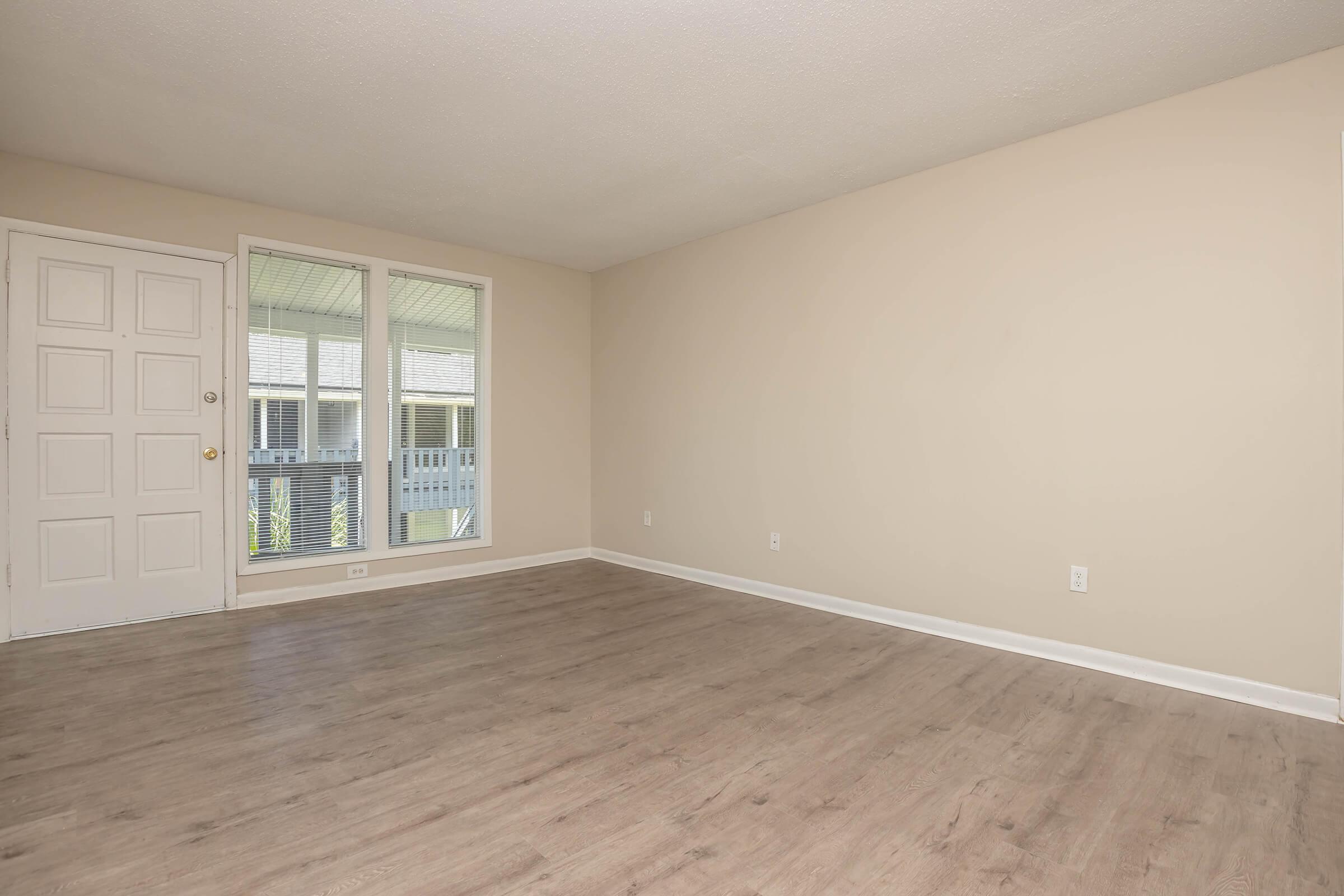 Spacious empty room with light beige walls and a large window allowing natural light. The floor is light-colored wood or laminate. A single door is visible on the left side, and there are no visible furnishings, creating a minimalist and open atmosphere.