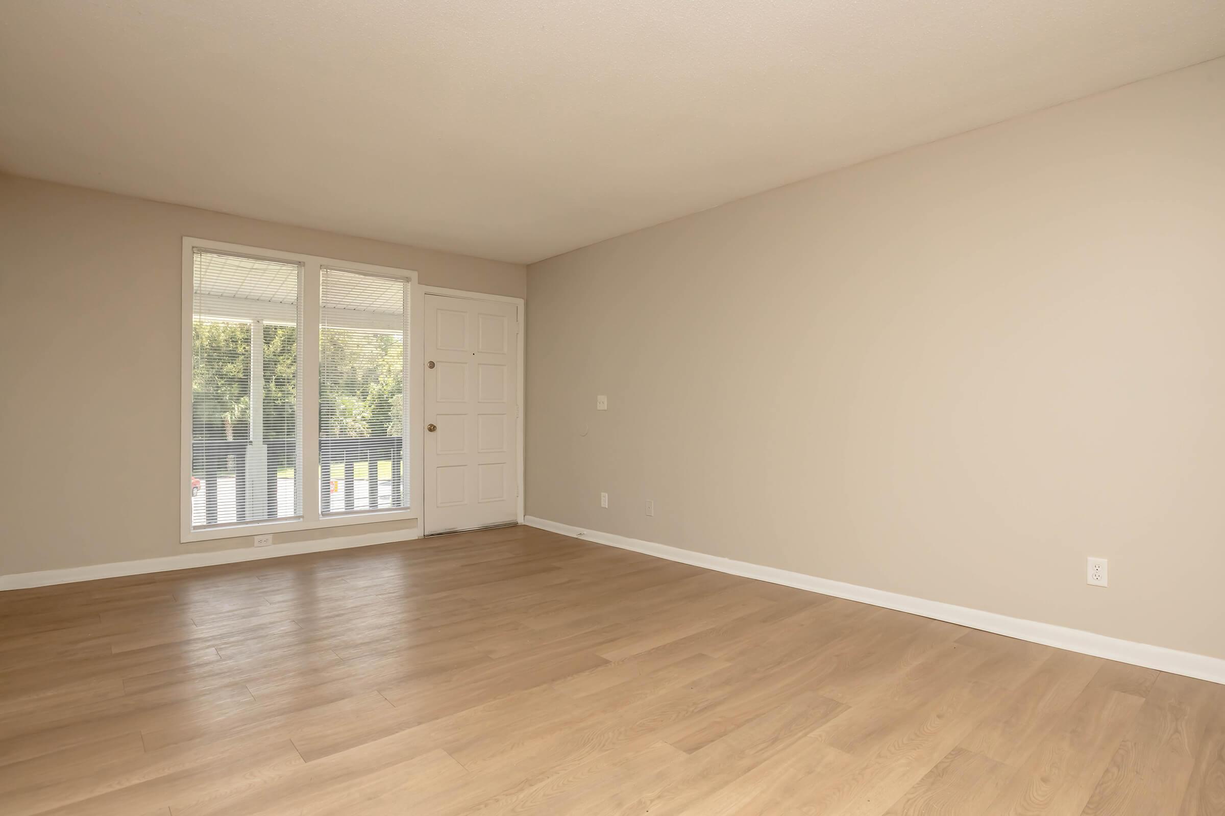 A spacious, unfurnished living room featuring light-colored walls and a wood floor. There are large windows providing natural light, and a front door with a simple design. The room has a clean and modern look, suitable for decorating.