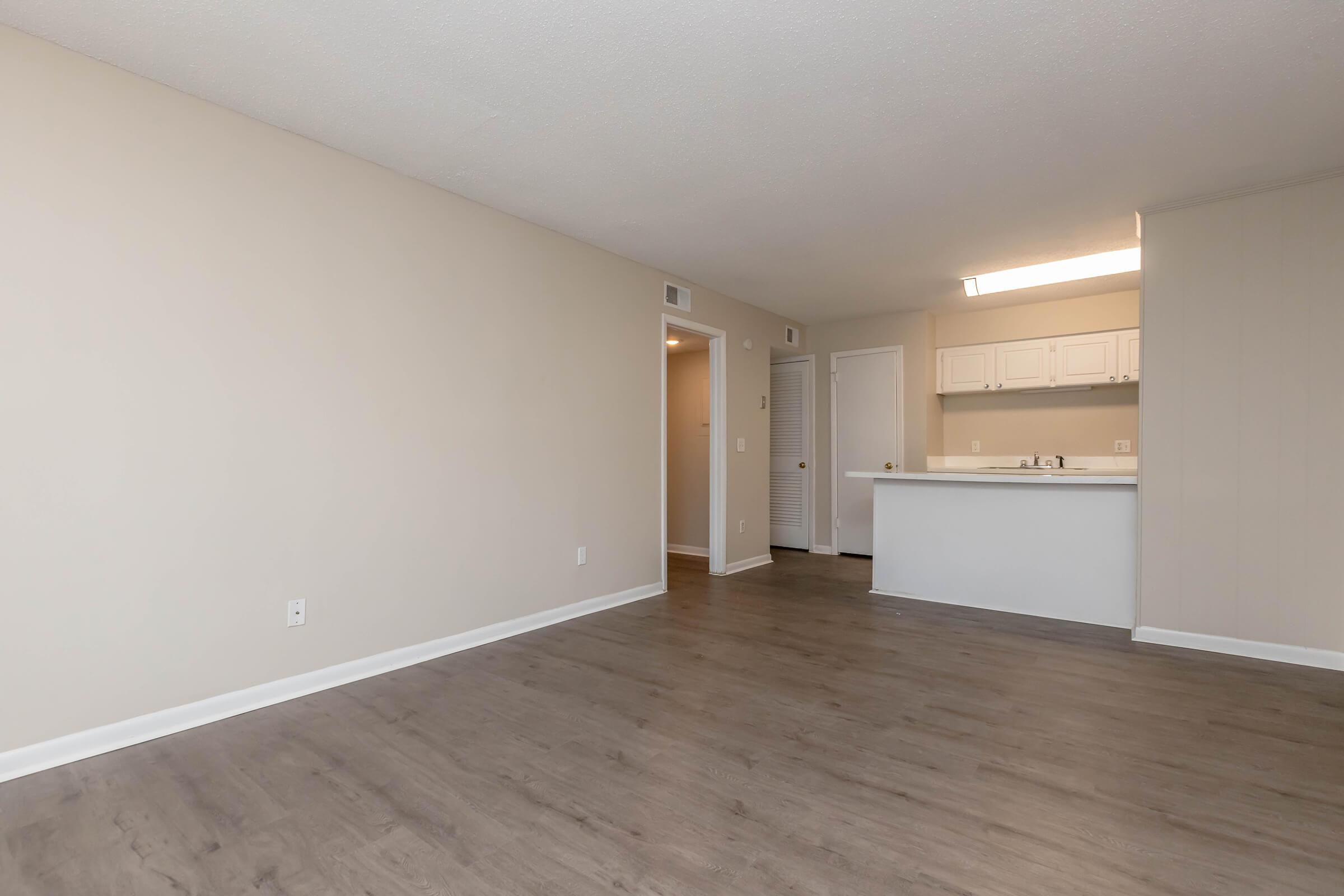 A spacious, empty living area with light-colored walls and a wooden floor. There’s a doorway leading to another room and an open kitchen with white cabinetry in the background. The lighting is bright, enhancing the airy feel of the space.