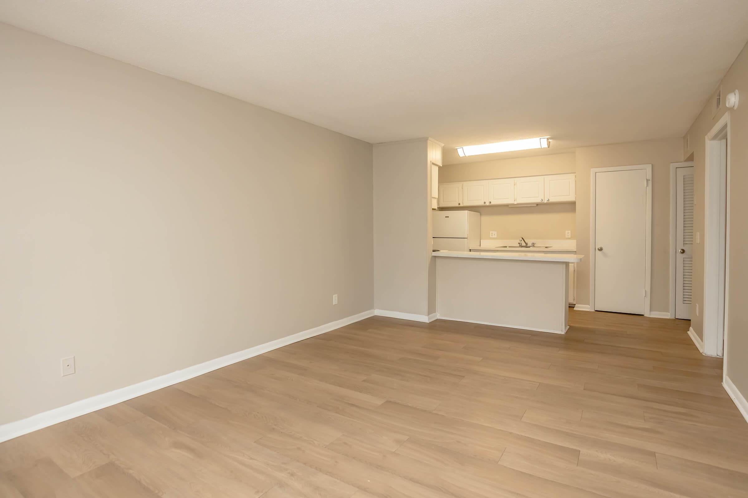 Empty room with light-colored walls and hardwood flooring, showcasing a spacious layout. In the background, a kitchen area is partially visible, featuring white cabinetry and appliances. Natural light fills the space, creating a bright and open atmosphere.