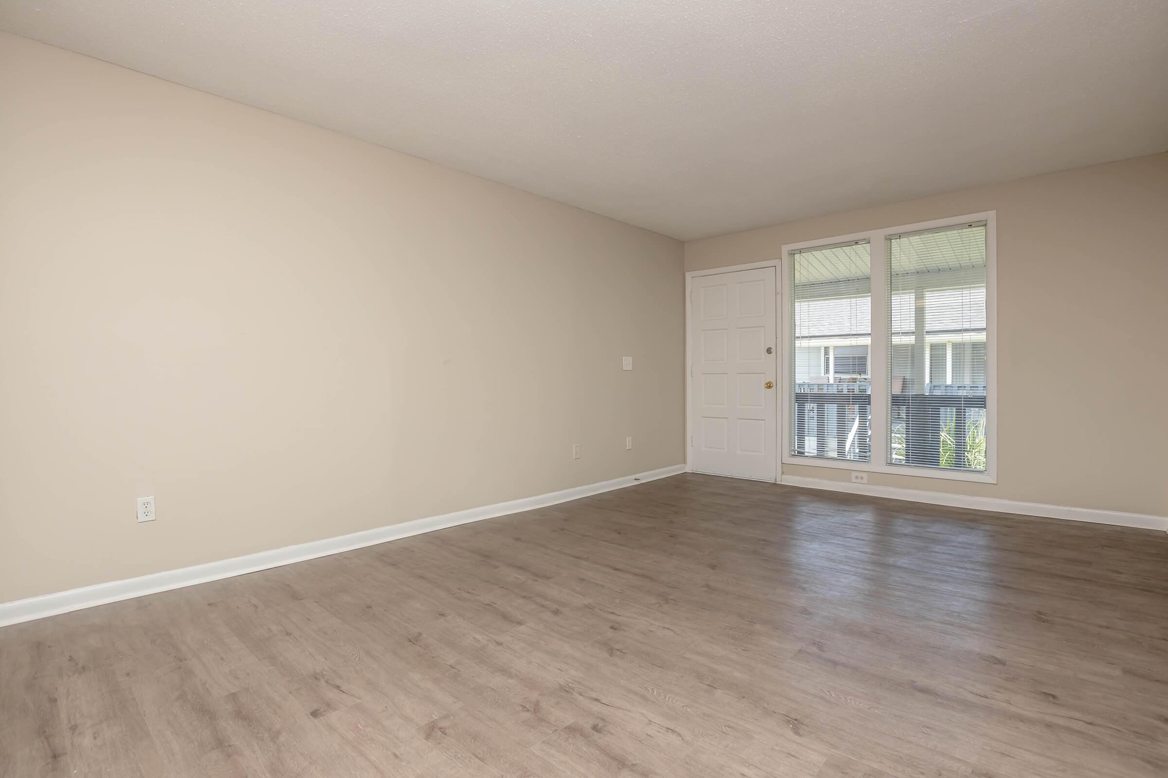 Empty living room featuring light-colored walls and a neutral wood-like floor. A single door is visible on the right, leading outside, and there are large windows allowing natural light in. The space is minimalistic and ready for furniture arrangement.