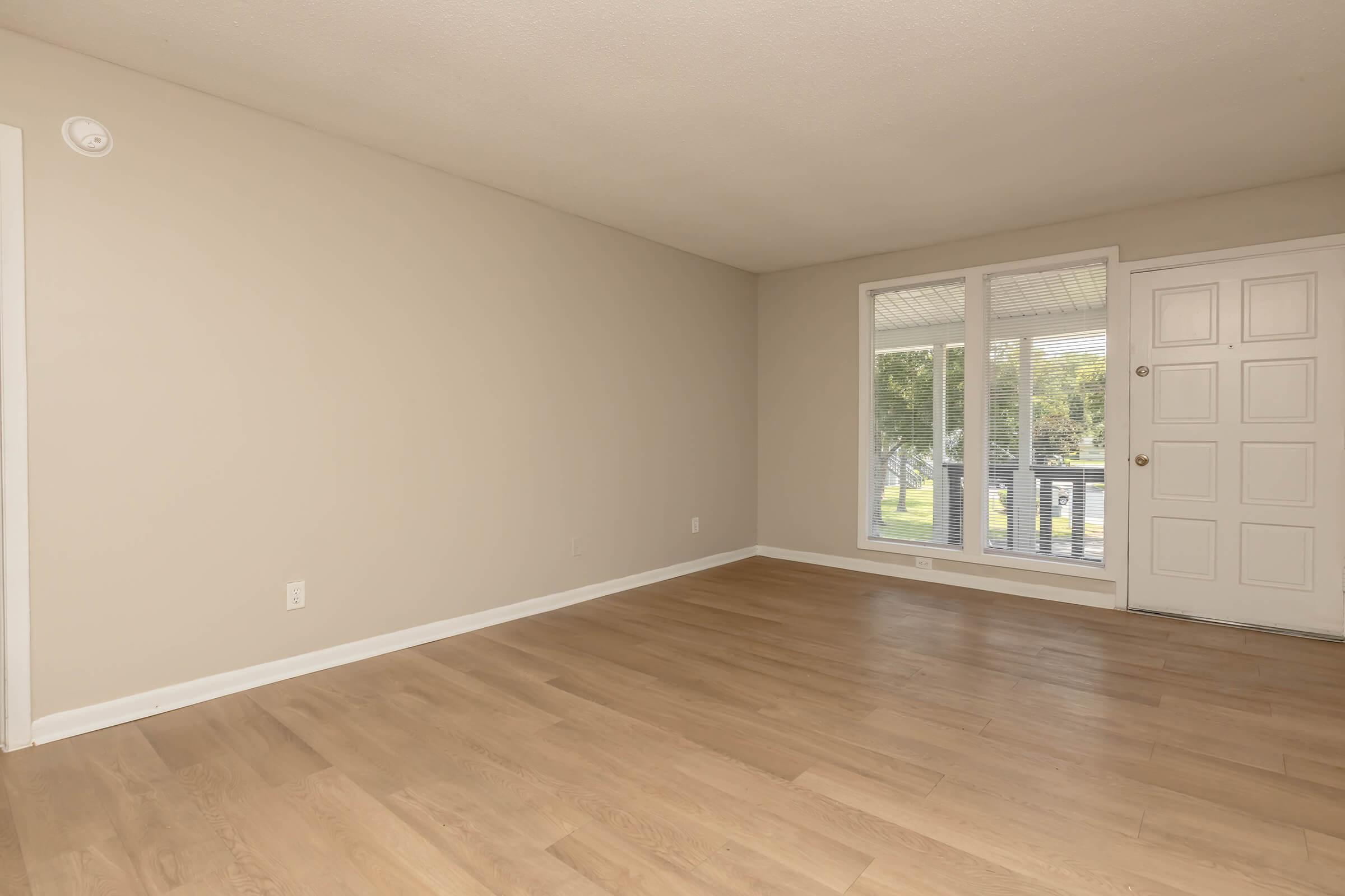 A spacious, empty living room with light-colored walls and wooden flooring. A large window with sheer curtains allows natural light to enter the room, while a front door is visible on the right side. The overall atmosphere is bright and airy, suitable for various interior design possibilities.