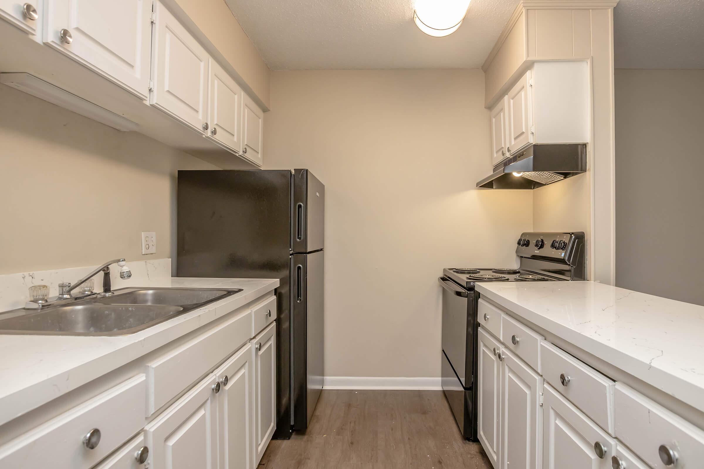 A modern kitchen featuring white cabinets, a stainless steel sink, a black refrigerator, and an electric stove with a hood. The countertops are light-colored, and the walls are a neutral beige. There's a light fixture on the ceiling, enhancing the space. The flooring is a wood laminate style.
