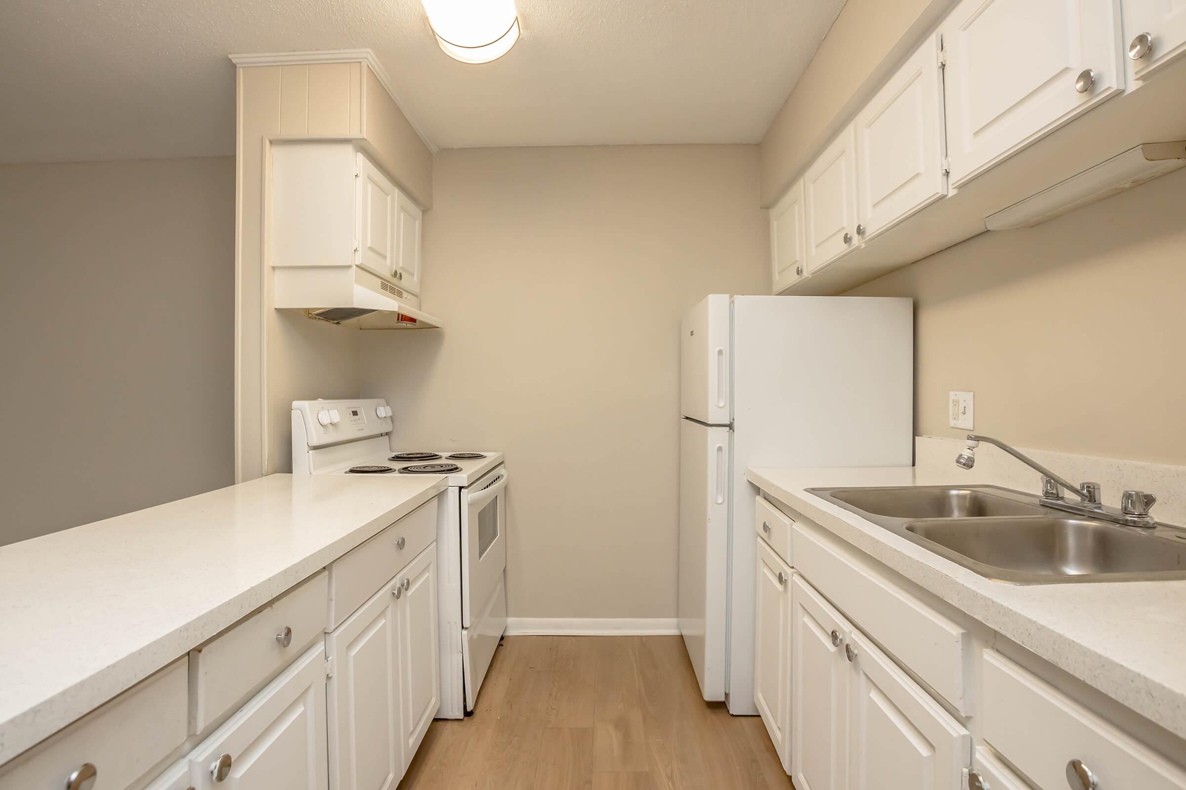 A modern kitchen featuring white cabinetry, a white refrigerator, an electric stove with an oven, and a double sink. The walls are painted in a light color, and the flooring is a natural wood finish. The space is well-lit by a ceiling light fixture.