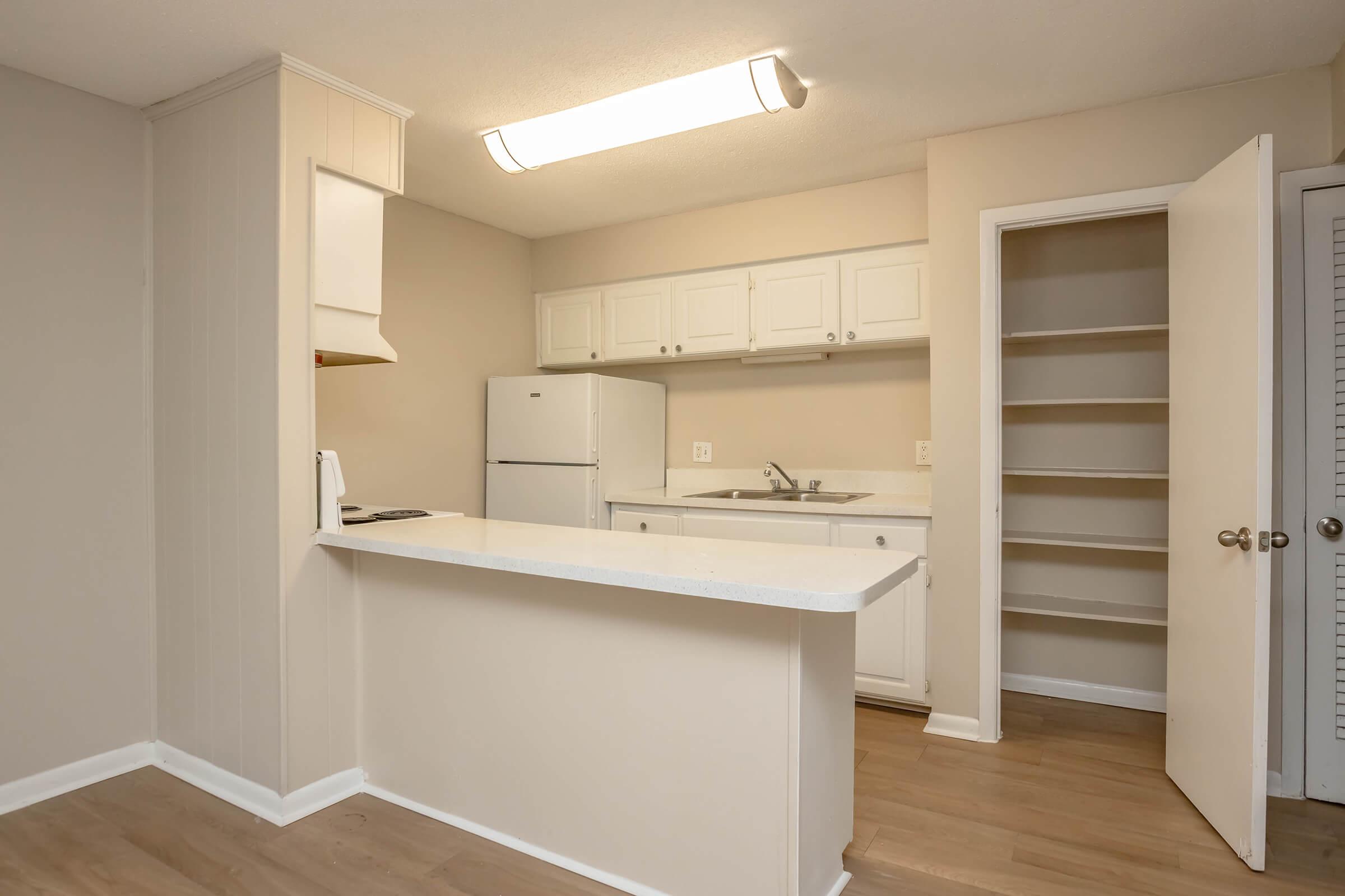 A clean and modern kitchen featuring white cabinetry, a refrigerator, and a sink. The kitchen has a small breakfast bar with countertop space, light-colored walls, and laminate flooring. A pantry is visible to the right, with an open door showcasing shelves. Natural light fills the space from a ceiling light fixture.
