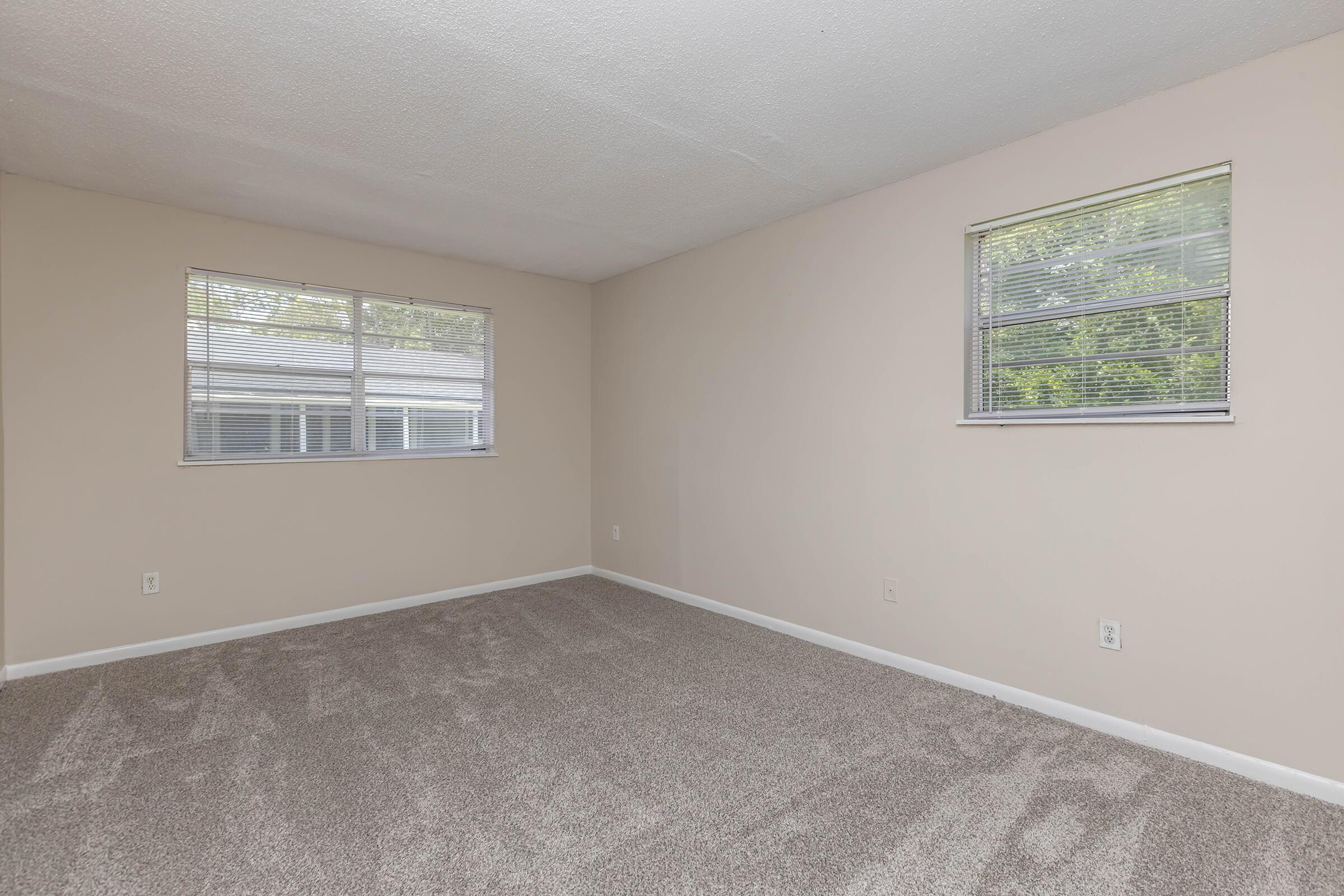 Spacious, empty room with light-colored walls and two windows allowing natural light. The floor is covered in gray carpet, and the overall ambiance is neutral and inviting, suitable for a variety of interior designs.