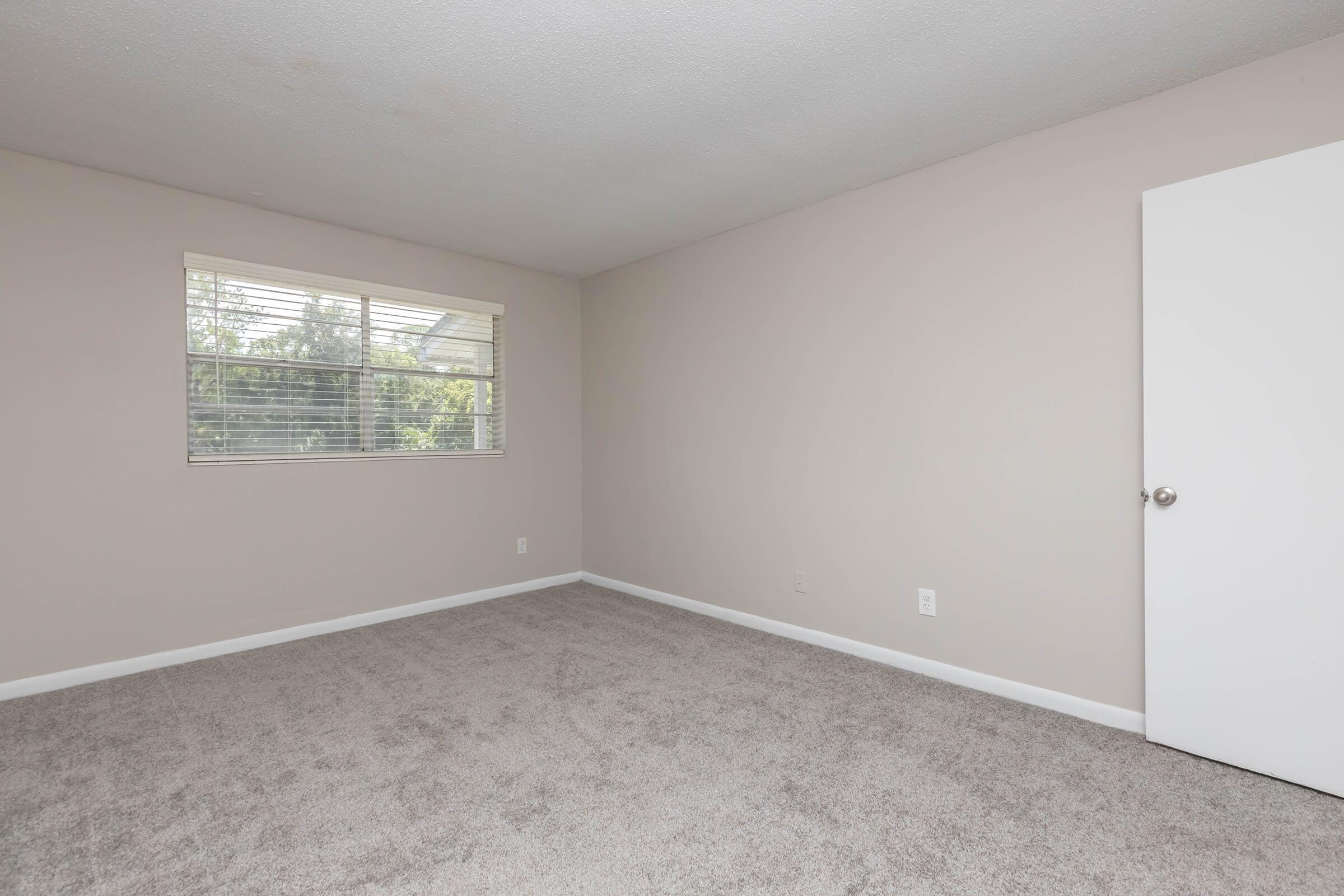 Empty room with light beige walls, a window allowing natural light, and gray carpet flooring. A white door is visible on the right. The space is uncluttered, offering a bright and airy atmosphere.