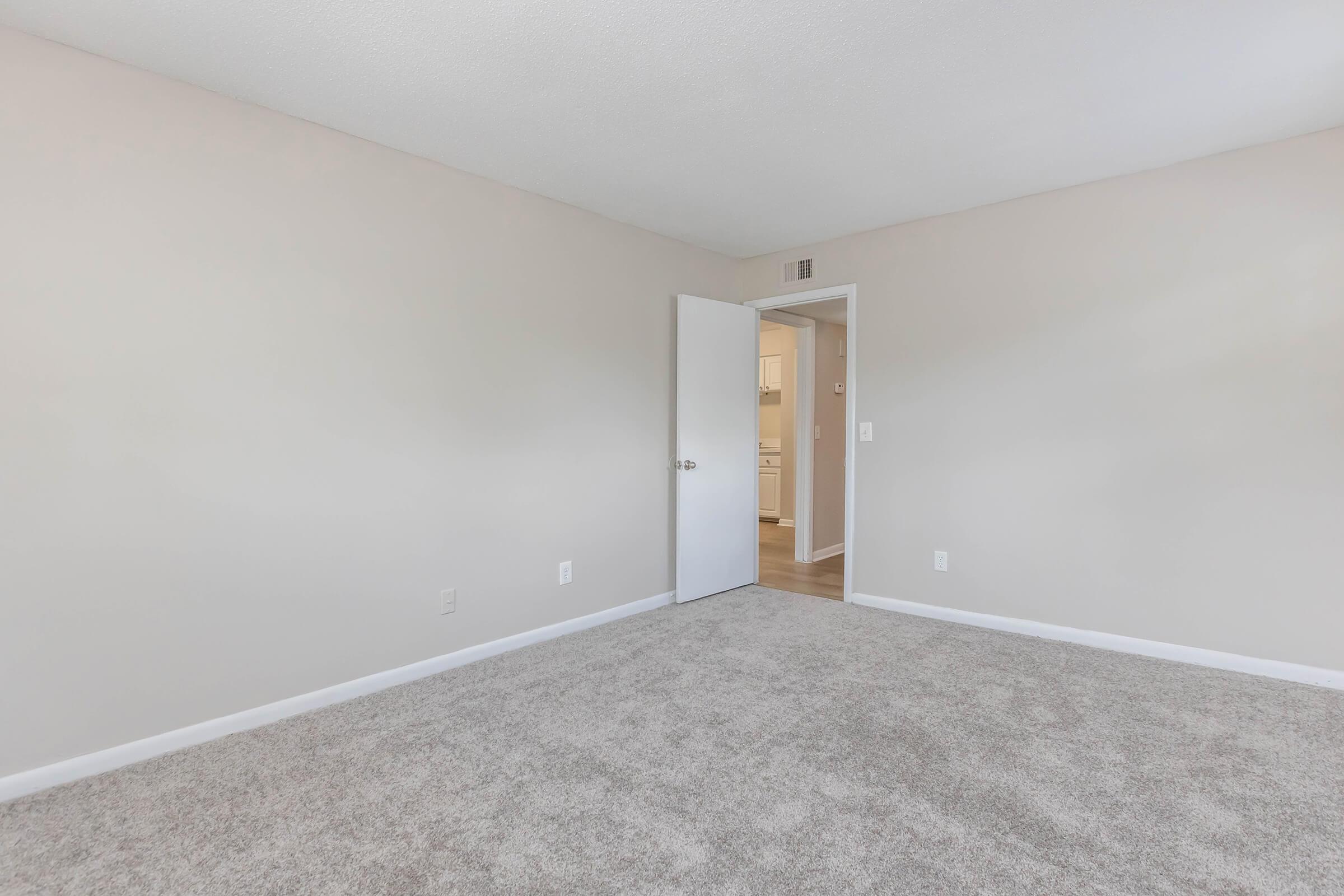 A vacant room with light beige walls and a plush carpet. A door on the left leads to another space, and the room has no furniture, creating an open and airy atmosphere. Natural light illuminates the area, enhancing the sense of spaciousness.