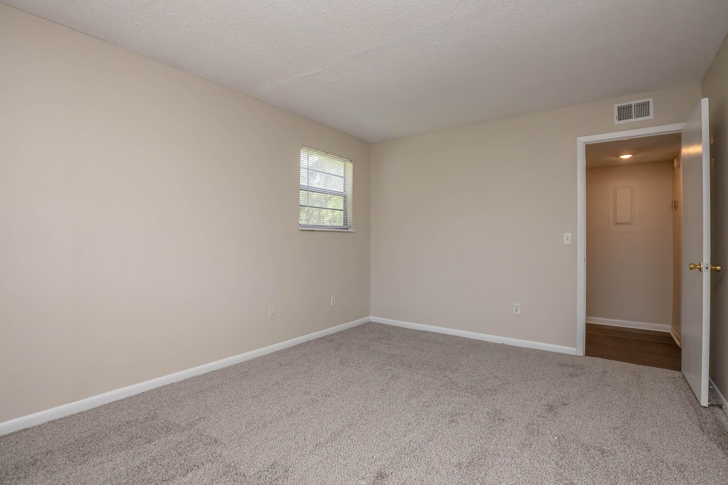 A spacious, empty room with light beige walls and a carpeted floor. There is a small window on the left side allowing natural light in. A door is visible on the right side, leading to another area. The room appears clean and ready for furnishing.