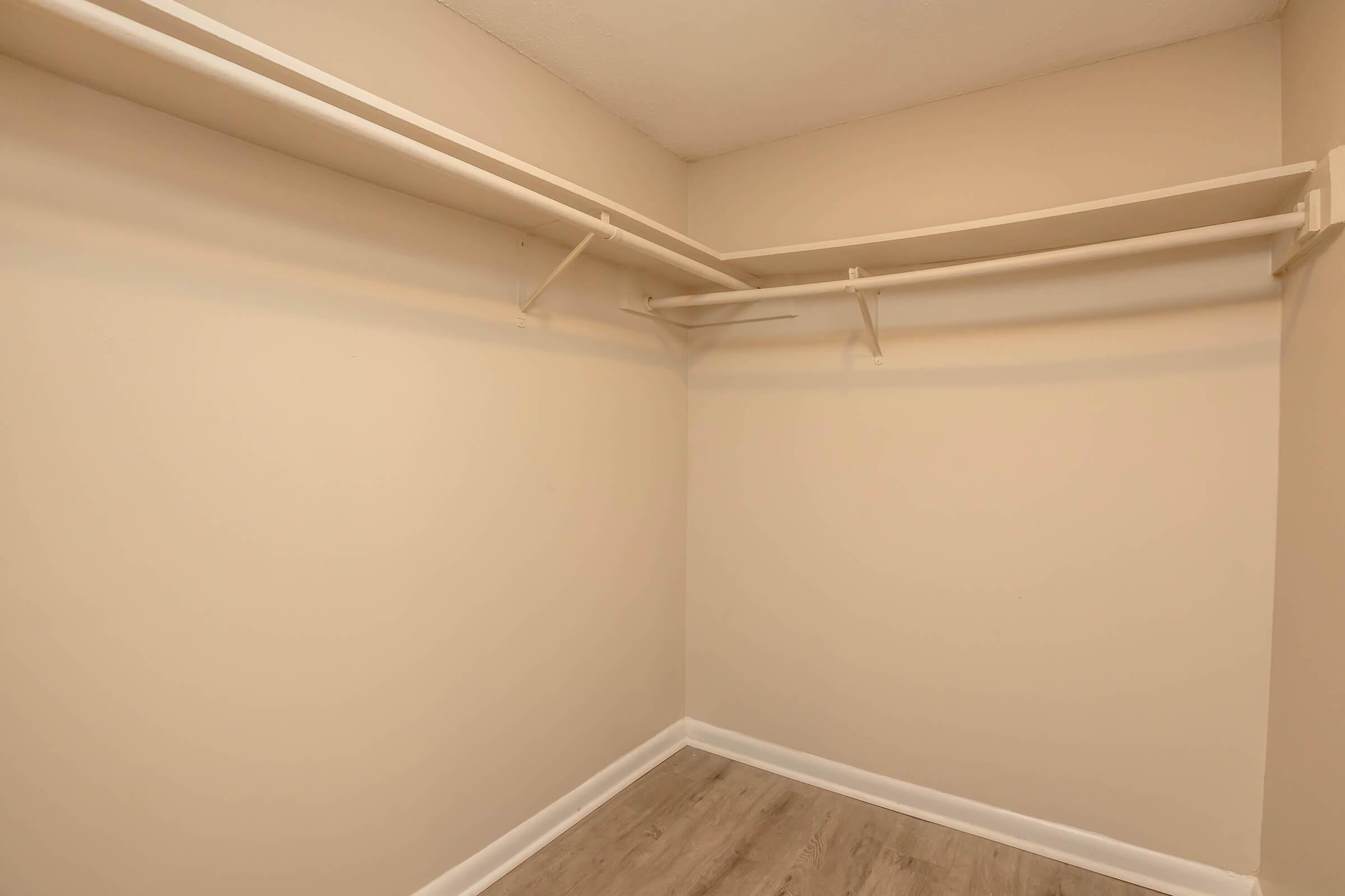 An empty closet with light beige walls and two white shelving units. The floor is covered with light wood laminate, and there are no items or decorations inside, creating a spacious and minimalist appearance.