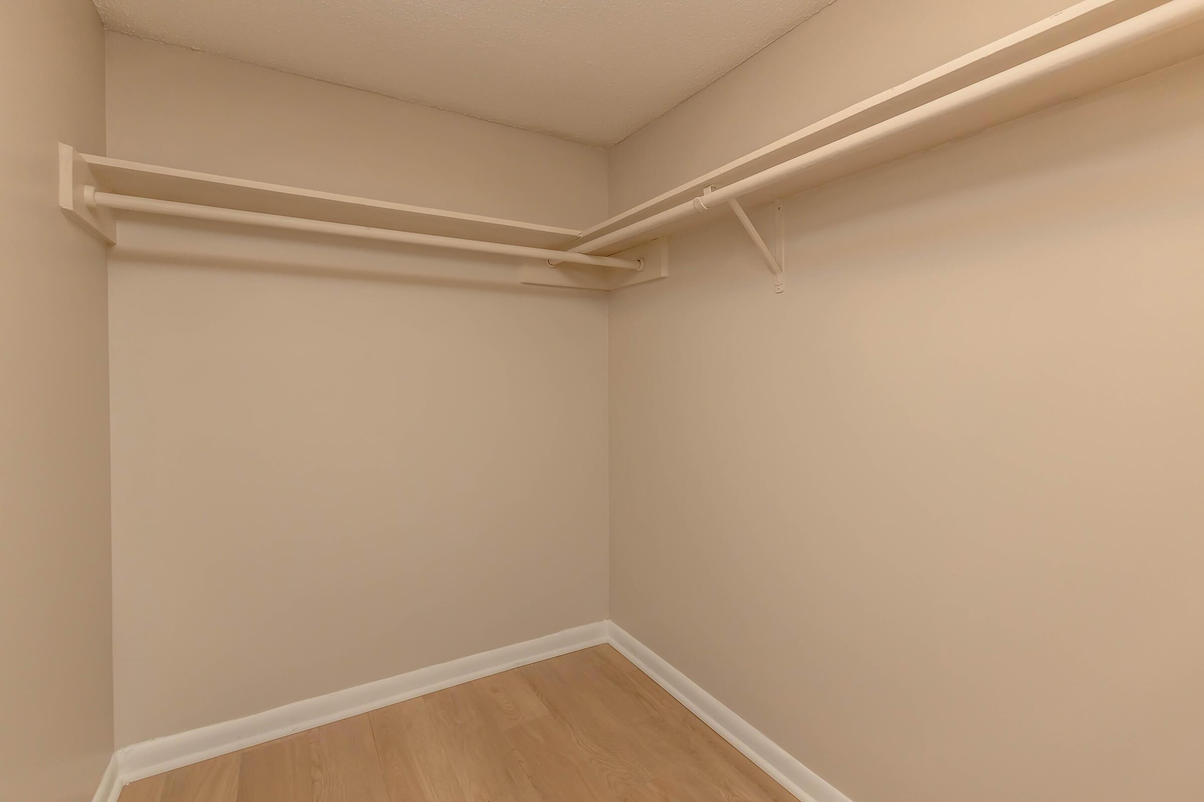 Empty closet space with light-colored walls and wooden flooring. The closet features two horizontal shelves positioned at different heights, providing potential for storage organization. The overall appearance is clean and minimalistic, ideal for various storage needs.