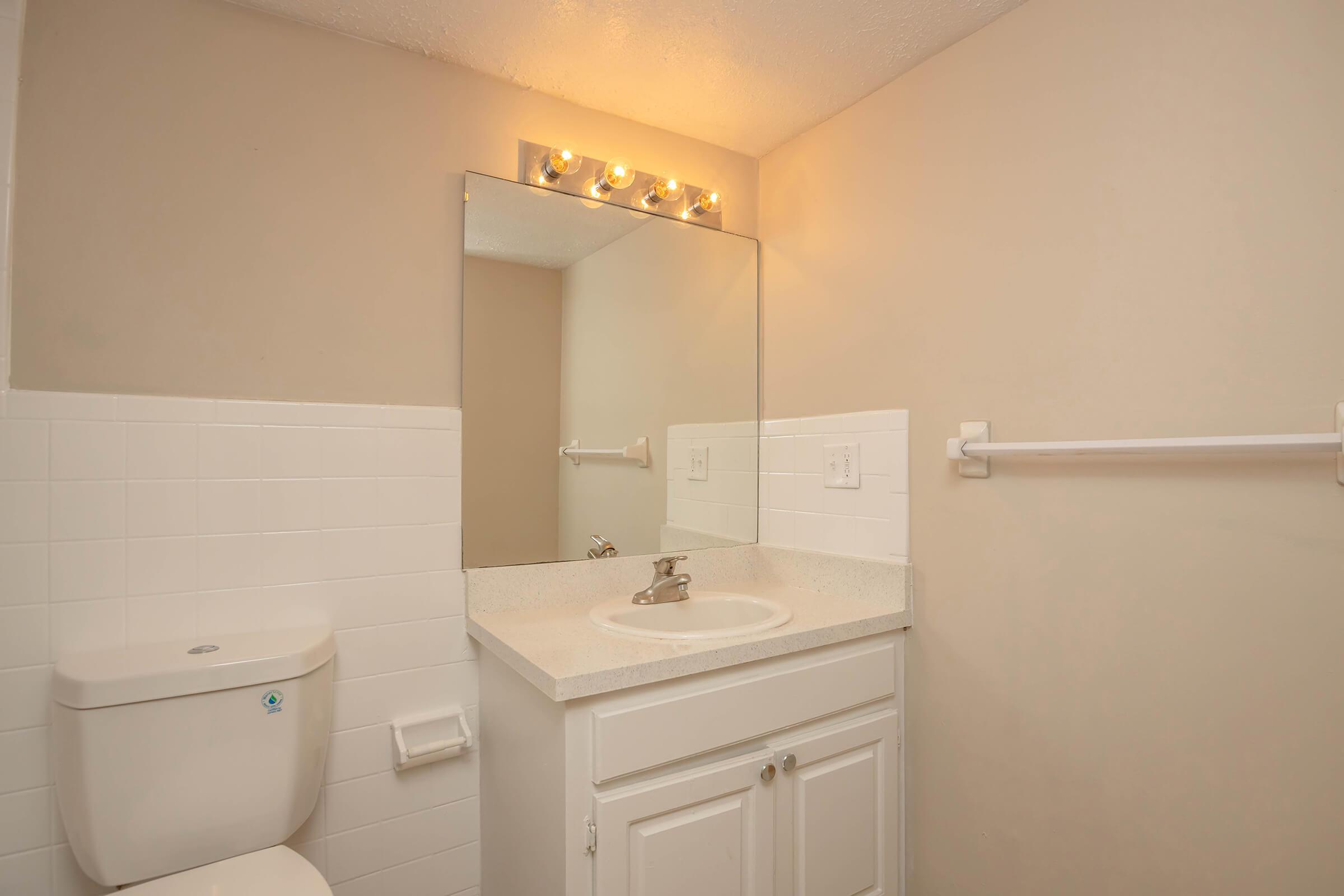 A clean bathroom featuring a wall-mounted mirror with overhead lights, a white sink with a faucet, a white toilet, and light-colored walls. The bathroom has a towel rack and tiled sections, creating a simple and modern look.