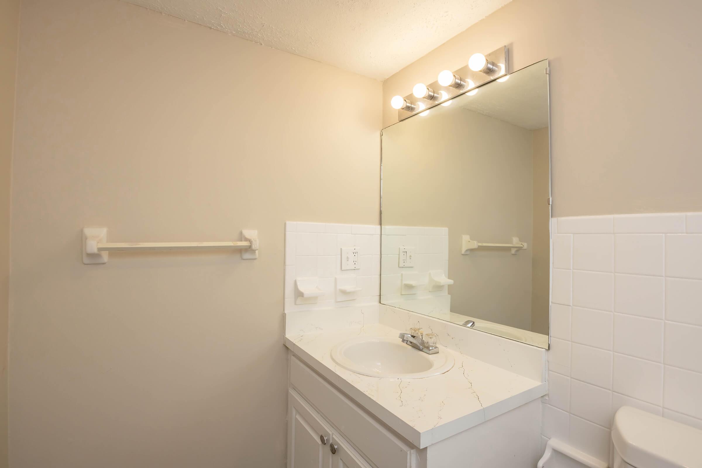 A clean, well-lit bathroom featuring a mirror with five light fixtures above, a white countertop sink, and a tiled wall. There are white towel racks and two soap dispensers mounted on the wall. The overall color scheme is neutral, with light beige walls and white fixtures.