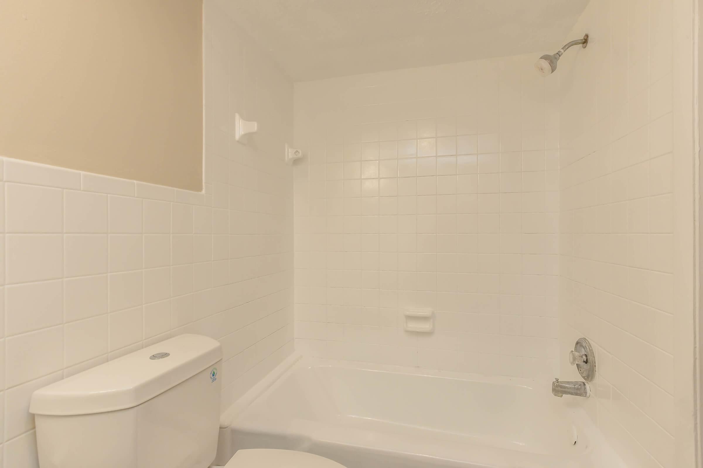 A clean, minimalist bathroom featuring a white tiled shower and bathtub, a white toilet, and a showerhead. The walls are also tiled in white, creating a bright and airy atmosphere. Natural light may be present, enhancing the space's cleanliness and simplicity.