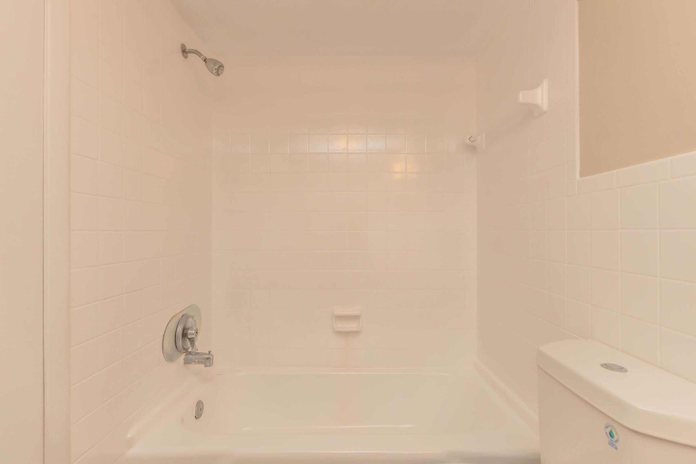 A clean, empty bathroom with a white tiled bathtub, a wall-mounted showerhead, a chrome faucet, and a toilet in the corner. The walls are painted in a light color, creating a bright and airy atmosphere.