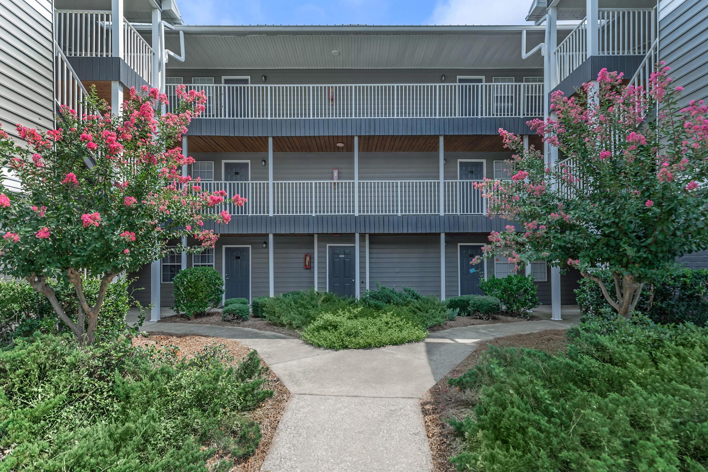 A well-maintained apartment building with three levels, featuring multiple balconies adorned with pink flowering plants in the foreground. The pathway leads through manicured shrubs and greenery, creating an inviting entrance. The building's exterior is clad in gray siding, with access doors visible along the front.