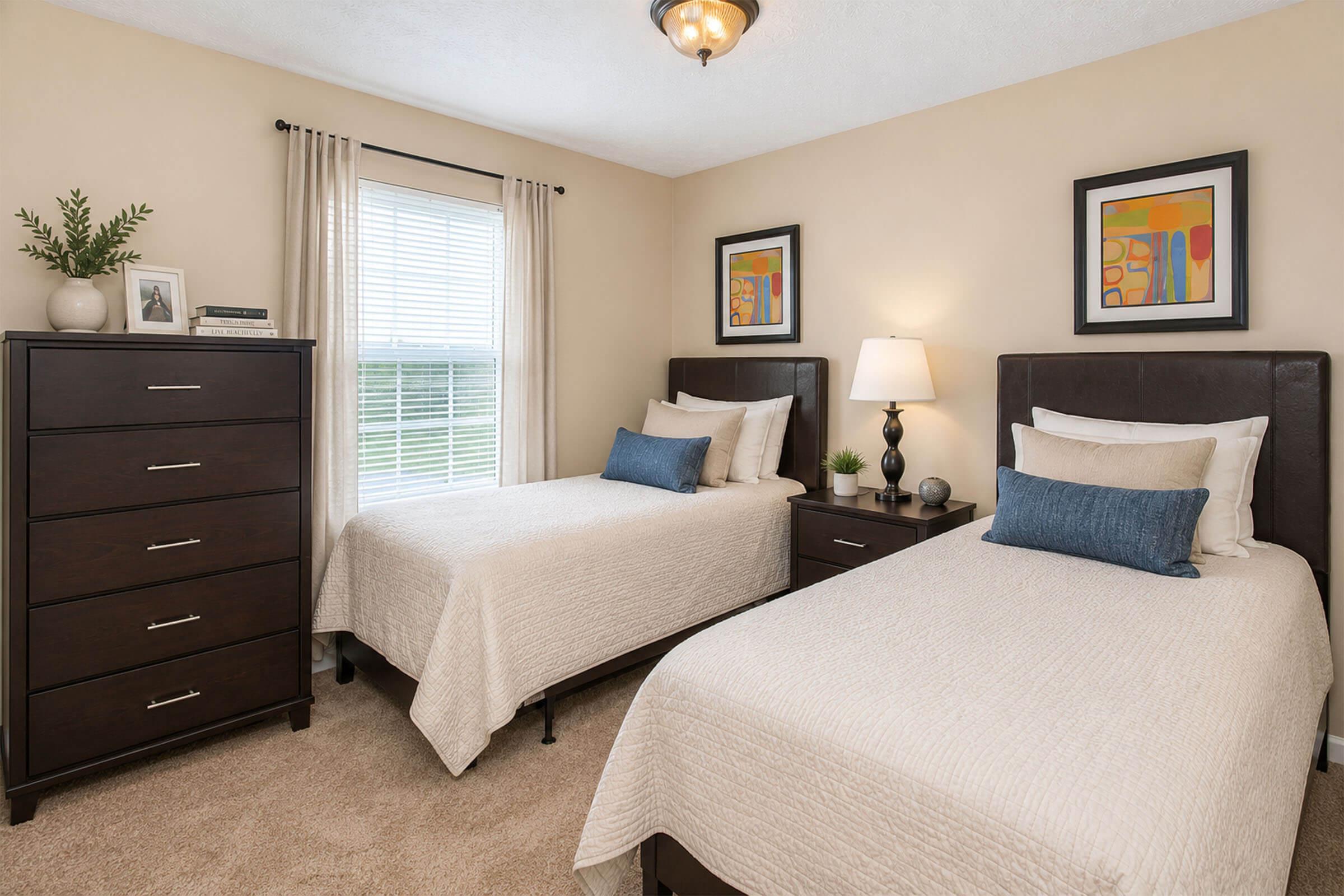 A well-decorated bedroom featuring two twin beds with neutral bedding, a bedside table with a lamp, and a dark wood dresser. The walls are painted a soft beige, and there are framed abstract artworks above each bed. A window allows natural light to fill the room, complemented by light curtains and a small plant on the dresser.