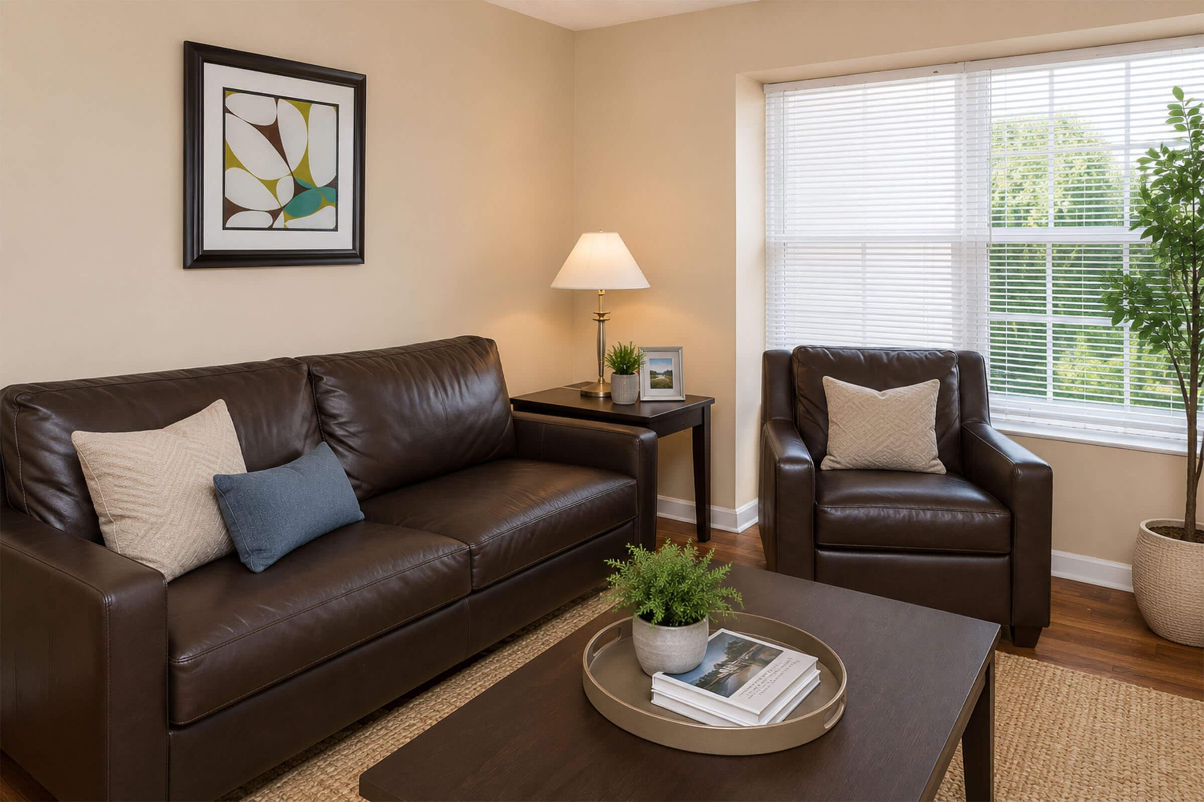 A cozy living room featuring a dark brown leather sofa with decorative pillows, a matching armchair, a wooden coffee table with a tray and magazine, a lamp on a side table, and a large window with blinds letting in natural light. A potted plant adds a touch of greenery to the space.