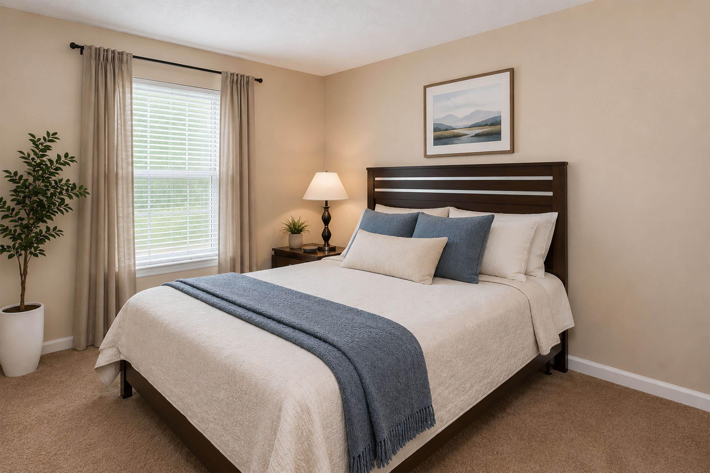 A cozy bedroom featuring a neatly made bed with a light-colored quilt and decorative pillows in shades of blue. There are bedside tables with lamps on each side. A window with beige curtains allows natural light in, and a potted plant adds a touch of greenery. A landscape painting hangs above the bed, complementing the warm decor.
