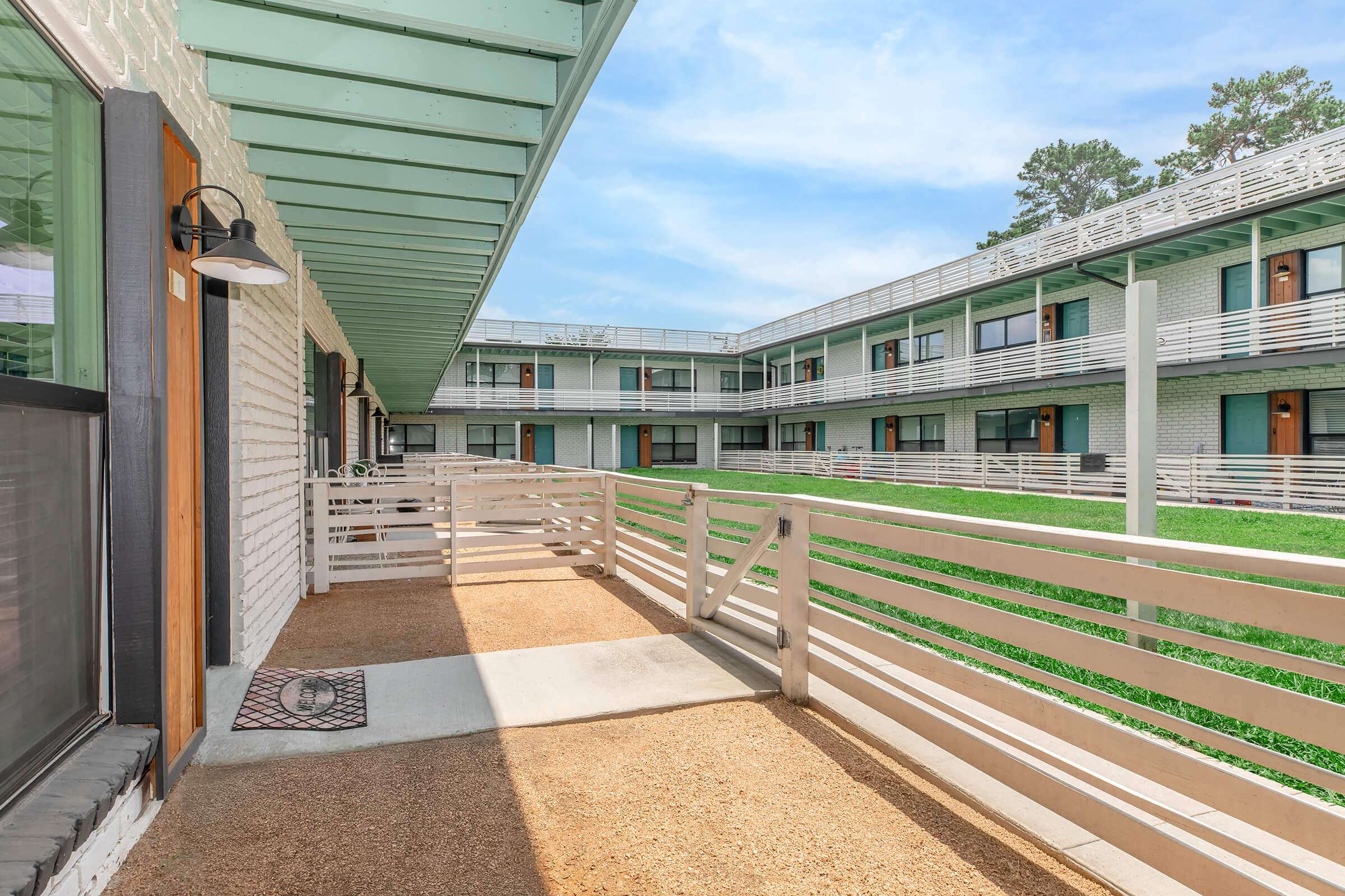 View of a well-lit hotel corridor with light green wooden overhangs. The walkway features a smooth surface leading to a grassy courtyard. Multiple doors line the corridor, opening into individual rooms, creating a spacious and inviting atmosphere. The sky is clear with a few clouds.