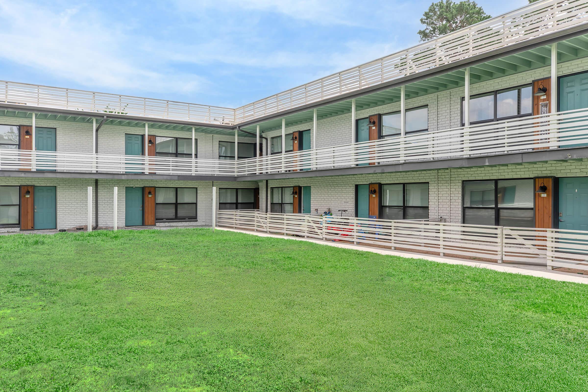 Two-story apartment complex with green grass in the foreground. The building features multiple entrances with blue doors and white railings on the balconies. Large windows are visible, providing an inviting look. The background has a clear blue sky, enhancing the overall bright and open atmosphere of the scene.