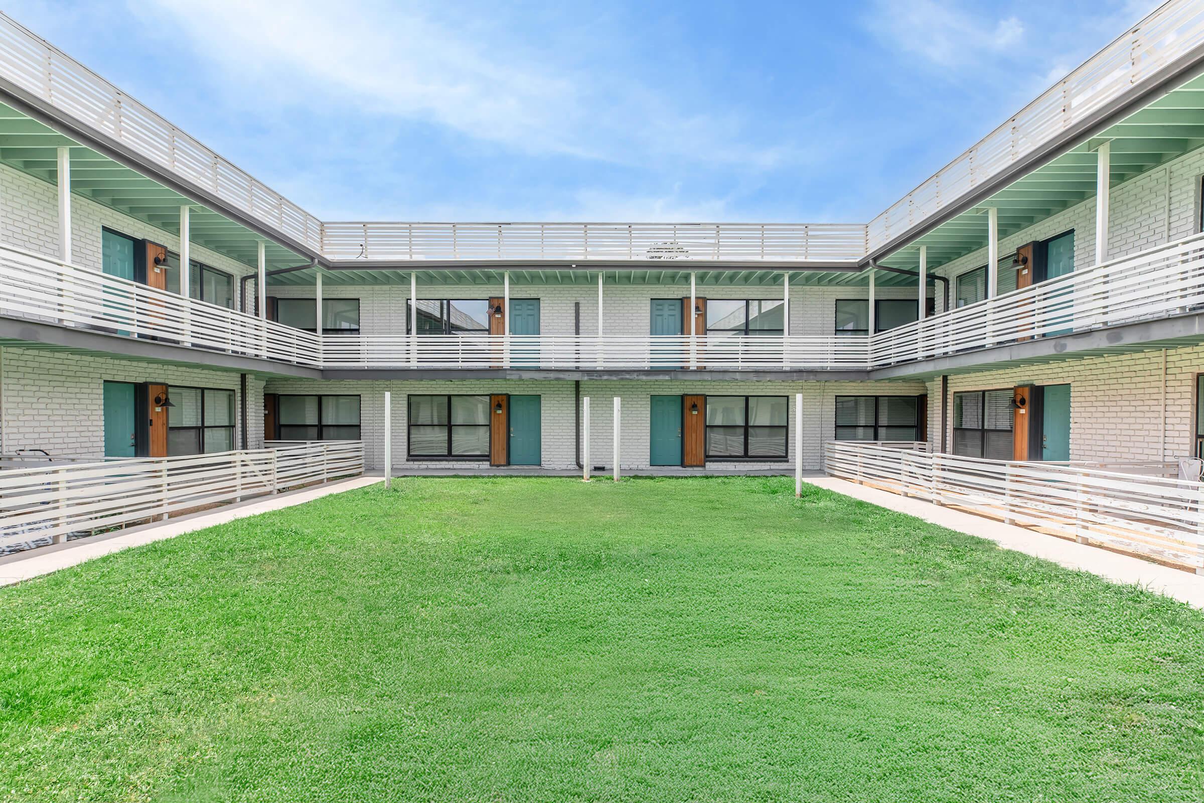 A well-maintained, two-story motel or apartment complex with a central grassy courtyard. The building features a light-colored exterior, multiple doorways, and balconies with white railings. Blue skies above complement the scene, creating a welcoming atmosphere.