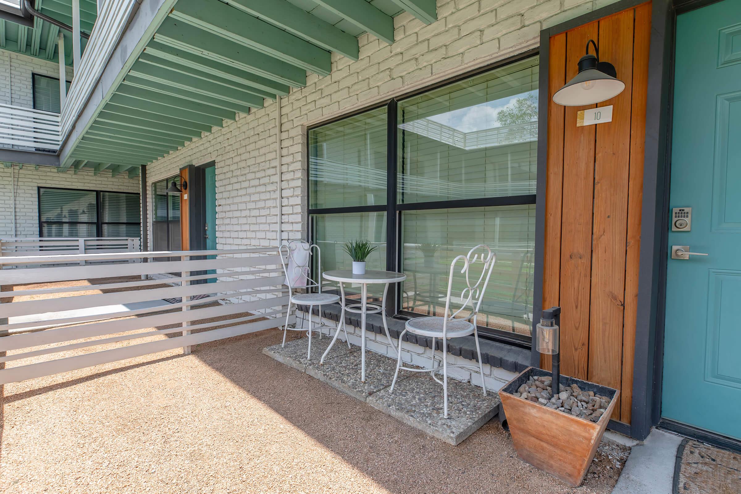 A cozy outdoor seating area features a round white table and two matching chairs on a gravel path. In the background, a modern building with large windows can be seen. A planter with rocks sits beside the door, which is painted a vibrant blue, adding a pop of color to the neutral exterior.
