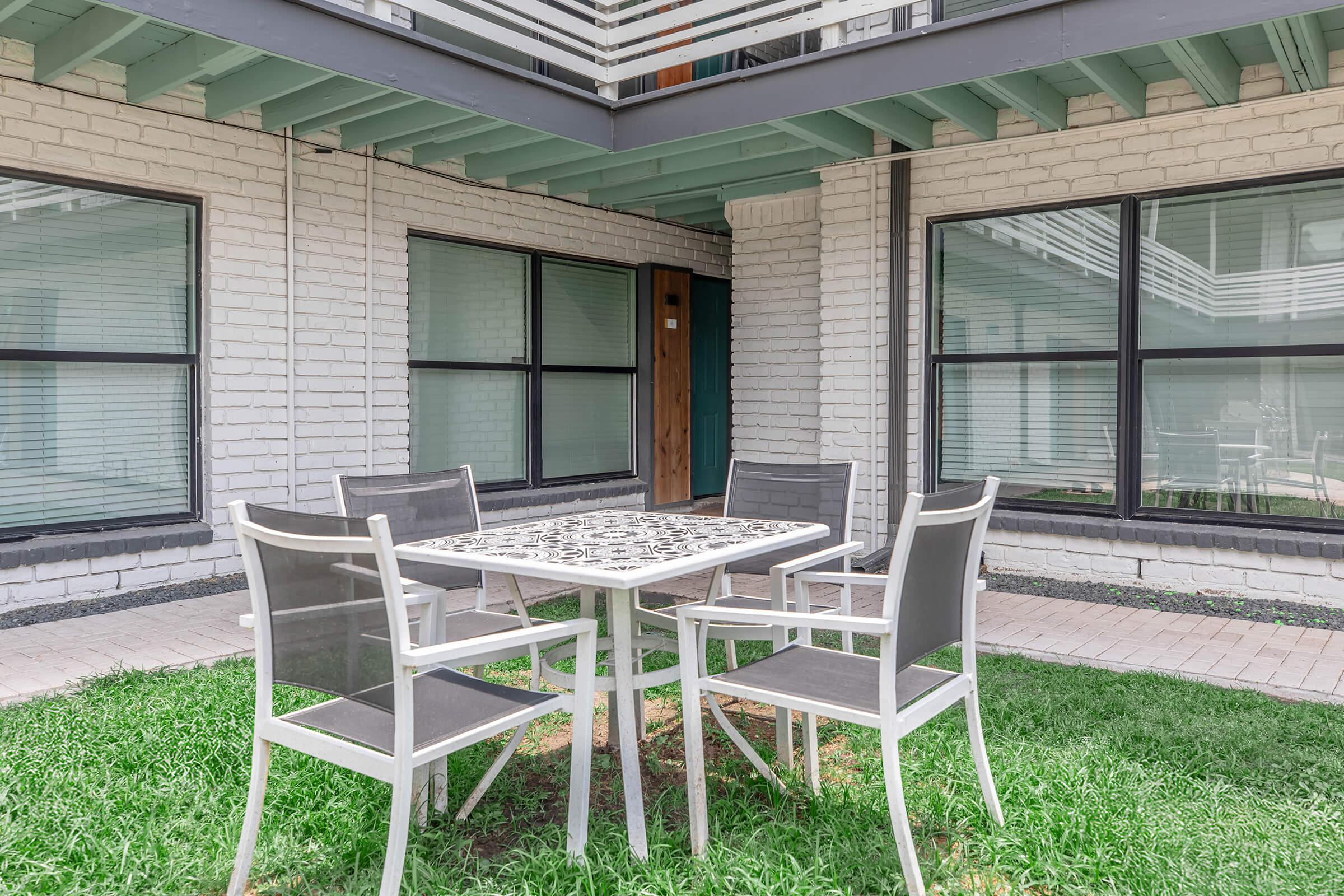 A patio area featuring a round table surrounded by four gray chairs, set on green grass. The background includes a white brick building with large windows and a wooden door, creating a contemporary outdoor space for relaxation.