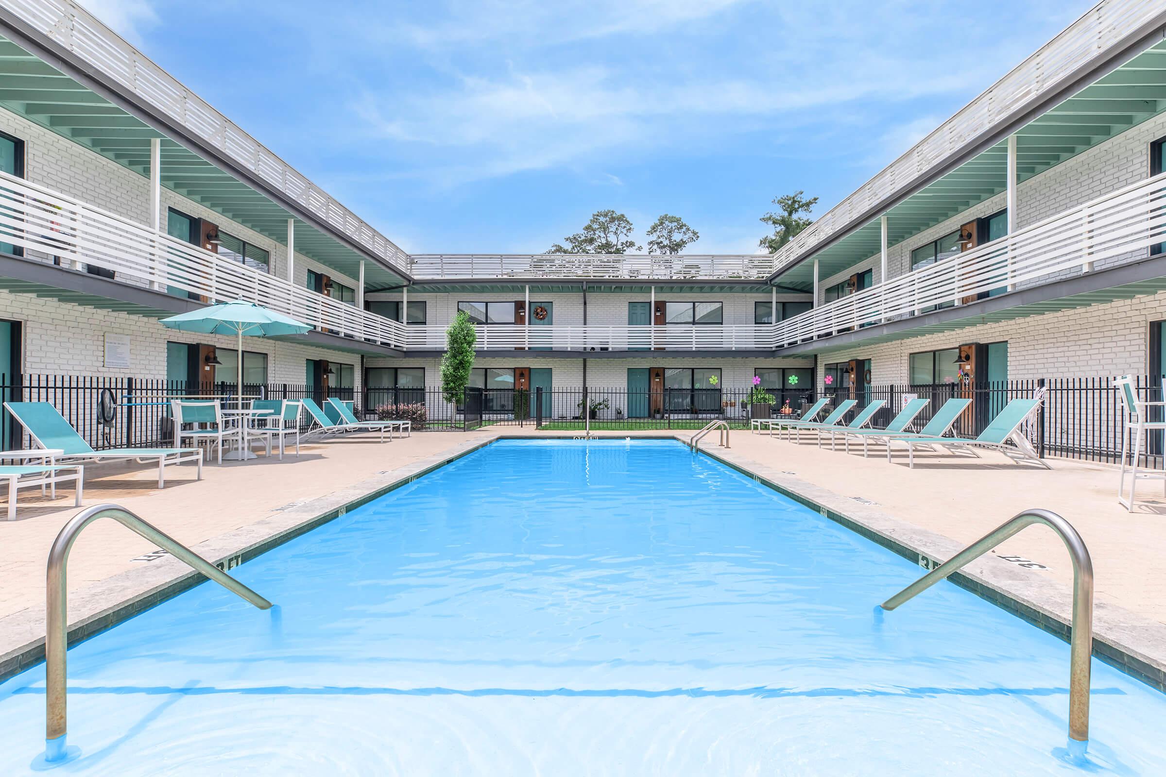 A well-maintained outdoor swimming pool surrounded by a courtyard featuring lounge chairs and umbrellas. The pool area is framed by a two-story building with multiple rooms. The sky is clear, and the setting is bright and inviting, suggesting a relaxing atmosphere.