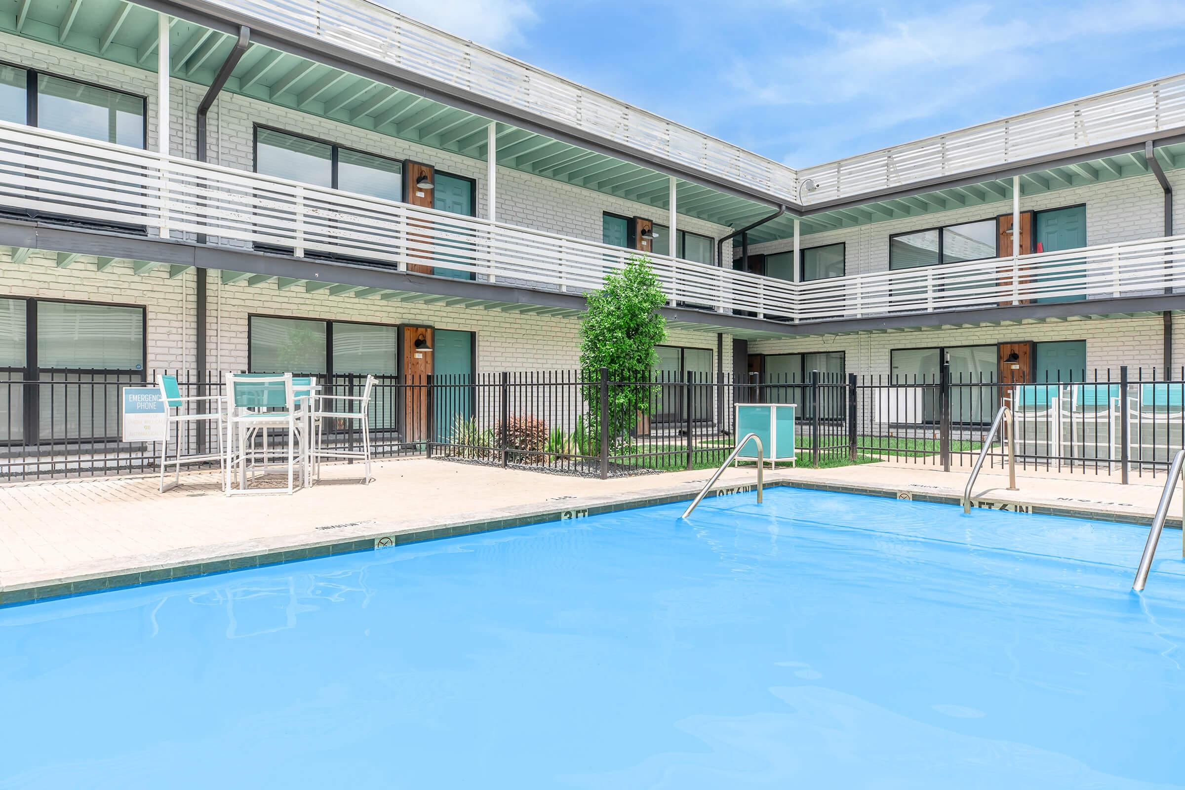 A clear blue swimming pool surrounded by a fenced area, with a modern hotel or apartment building in the background. The building features multiple balconies and large windows, and there are a few chairs positioned by the pool. The sky is bright blue with some clouds.