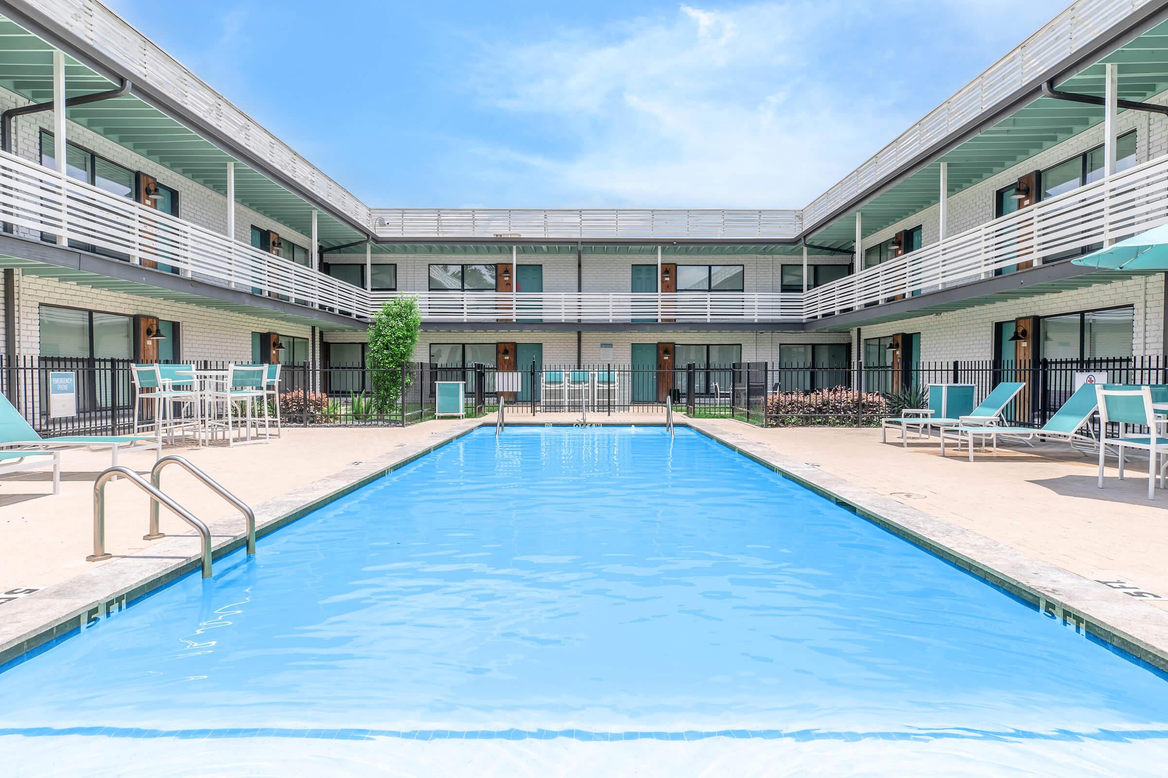 A clear blue swimming pool surrounded by lounge chairs and a patio area. Two stories of buildings with balconies overlook the pool. The scene is set in bright daylight, with trees and landscaping visible in the background, creating a relaxing atmosphere.