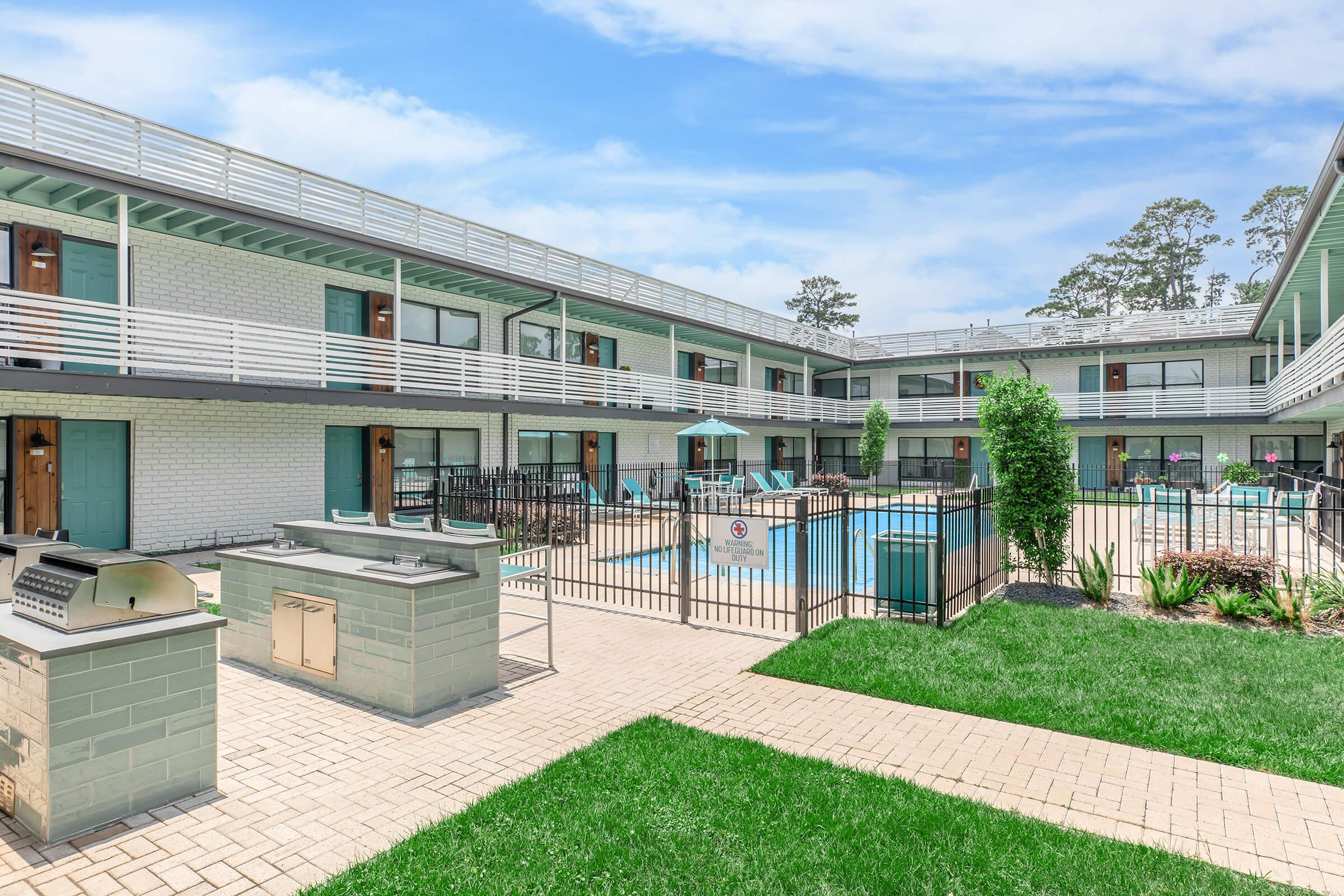A view of an outdoor area of a multi-story apartment complex featuring multiple units with balconies. In the center, there is a swimming pool surrounded by lounge chairs and a fence. There are grassy areas and a barbecue grill nearby, with trees in the background and a partly cloudy sky.