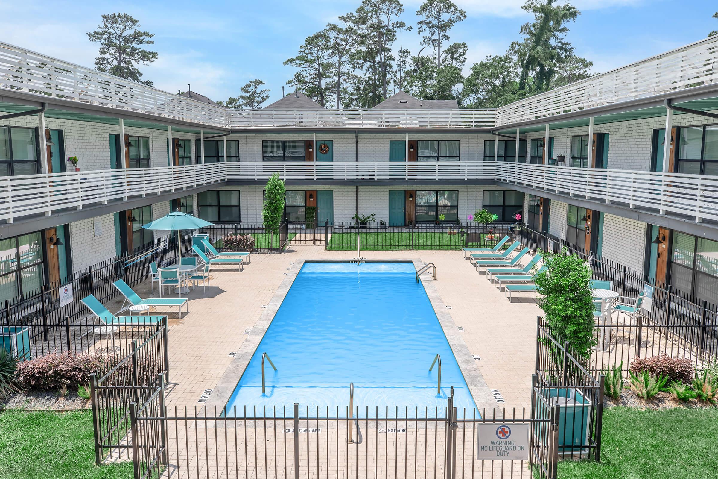 A view of a courtyard pool area surrounded by a two-story apartment building. The pool is clean and illuminated, with several lounge chairs and umbrellas arranged around it. Lush greenery and flowers enhance the area, and a black fence encloses the pool for safety.