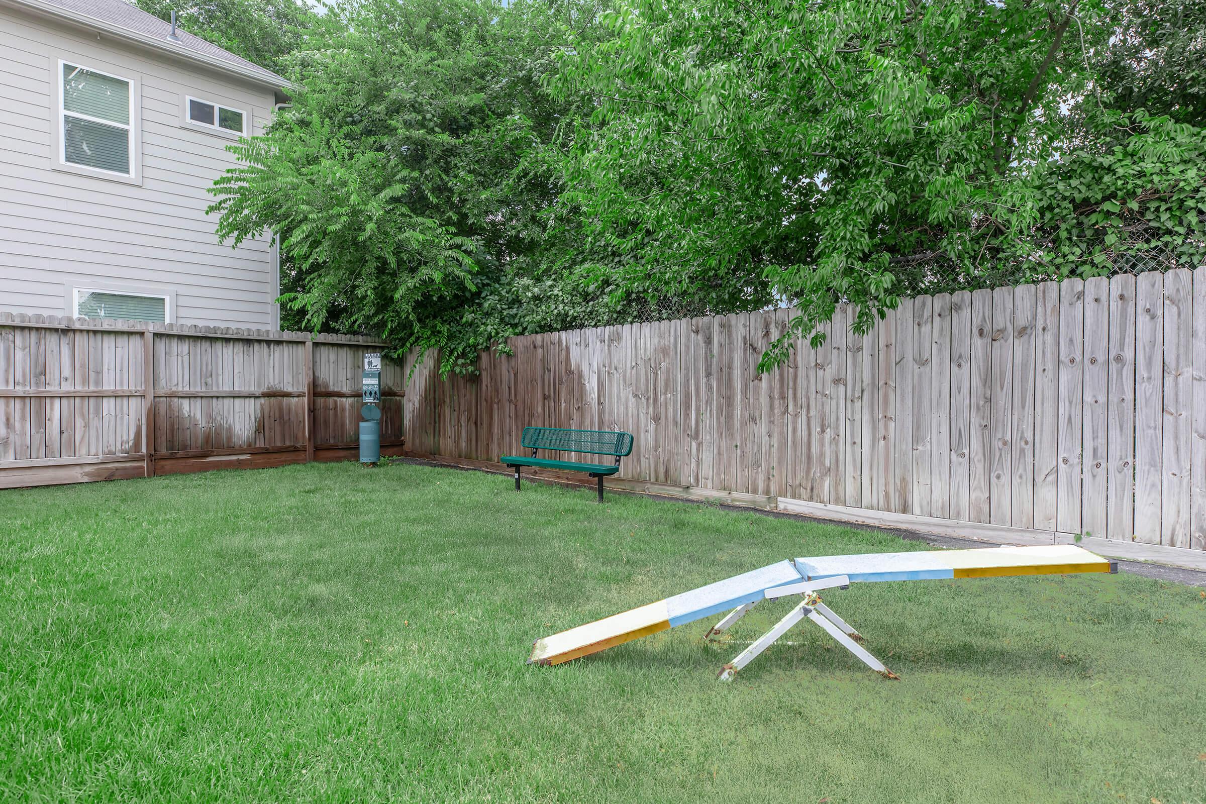 A grassy backyard featuring a wooden seesaw painted in bright colors, a green bench along the fence, and a wooden fence surrounding the area. Trees are visible in the background, adding greenery to the setting.