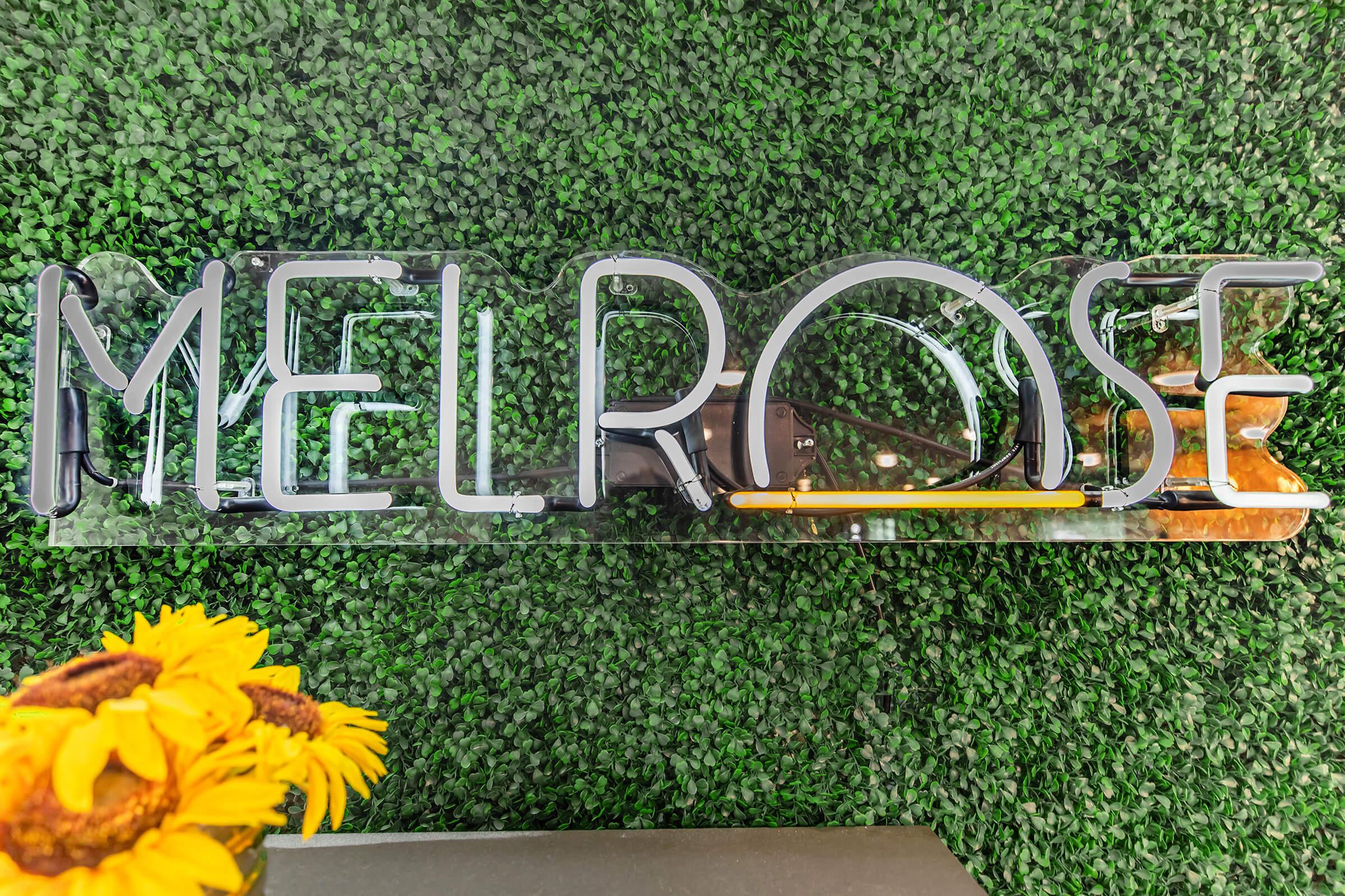 A clear sign displaying the word "MELROSE," set against a background of artificial green foliage. A small bouquet of sunflowers is partially visible in the lower left corner, adding a vibrant touch to the scene.