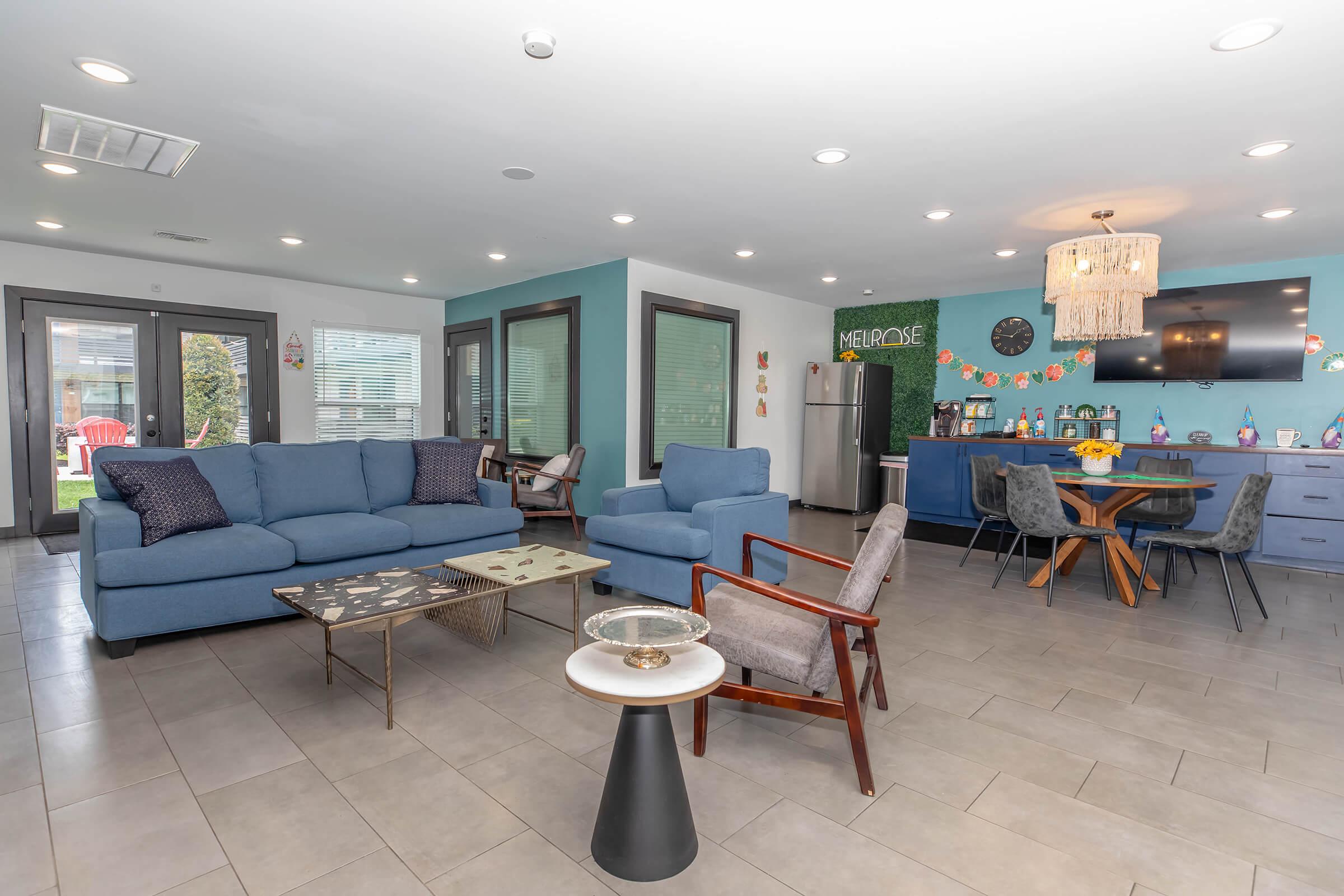 A modern, open-concept living space featuring blue sofas, a glass coffee table, and stylish dining chairs. The kitchen area is equipped with a refrigerator and has a decorative wall, while large windows allow natural light to brighten the room. A welcoming atmosphere is emphasized by decorative details.