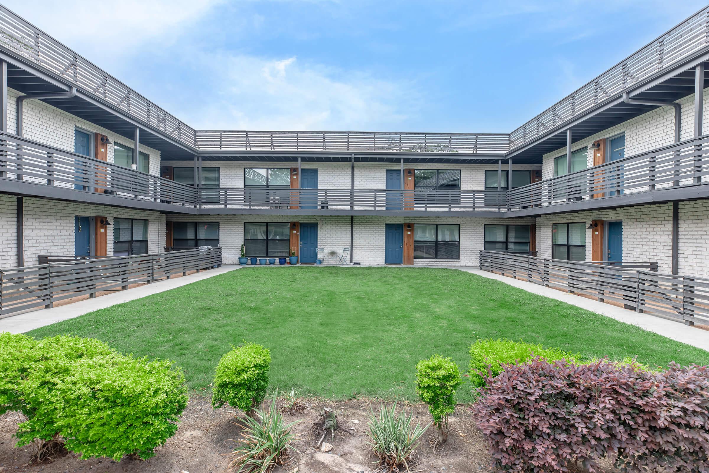 A courtyard view of a two-story apartment building featuring a grassy area and neatly trimmed shrubs. The building has a modern design with balconies, railing, and multiple doors leading to the apartments. The sky is clear, adding brightness to the scene.